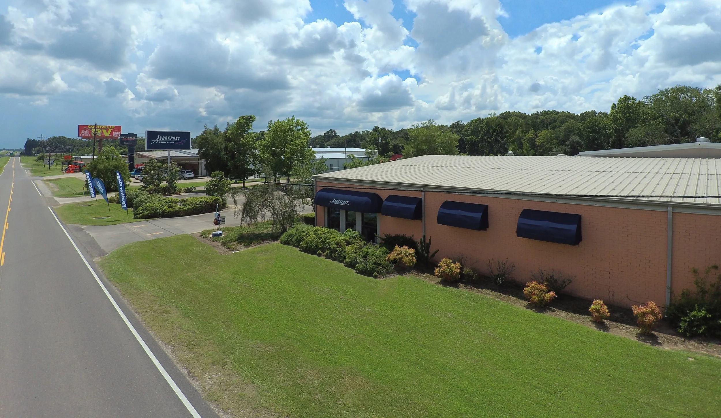 A commercial building with a brick exterior and three blue awnings, surrounded by landscaped bushes and grass, located near a road with a billboard and other businesses in the background, under partly cloudy skies.
