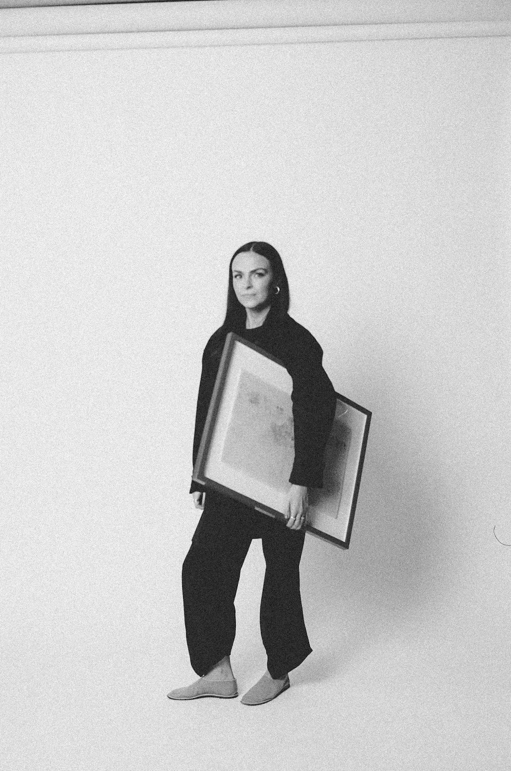 Founder of EMD Studio (Emily Mauldin Design Studio), Emily Mauldin, designer and creative, with dark hair standing against a plain light-colored background, holding two framed pictures, wearing dark clothing and light-colored shoes.