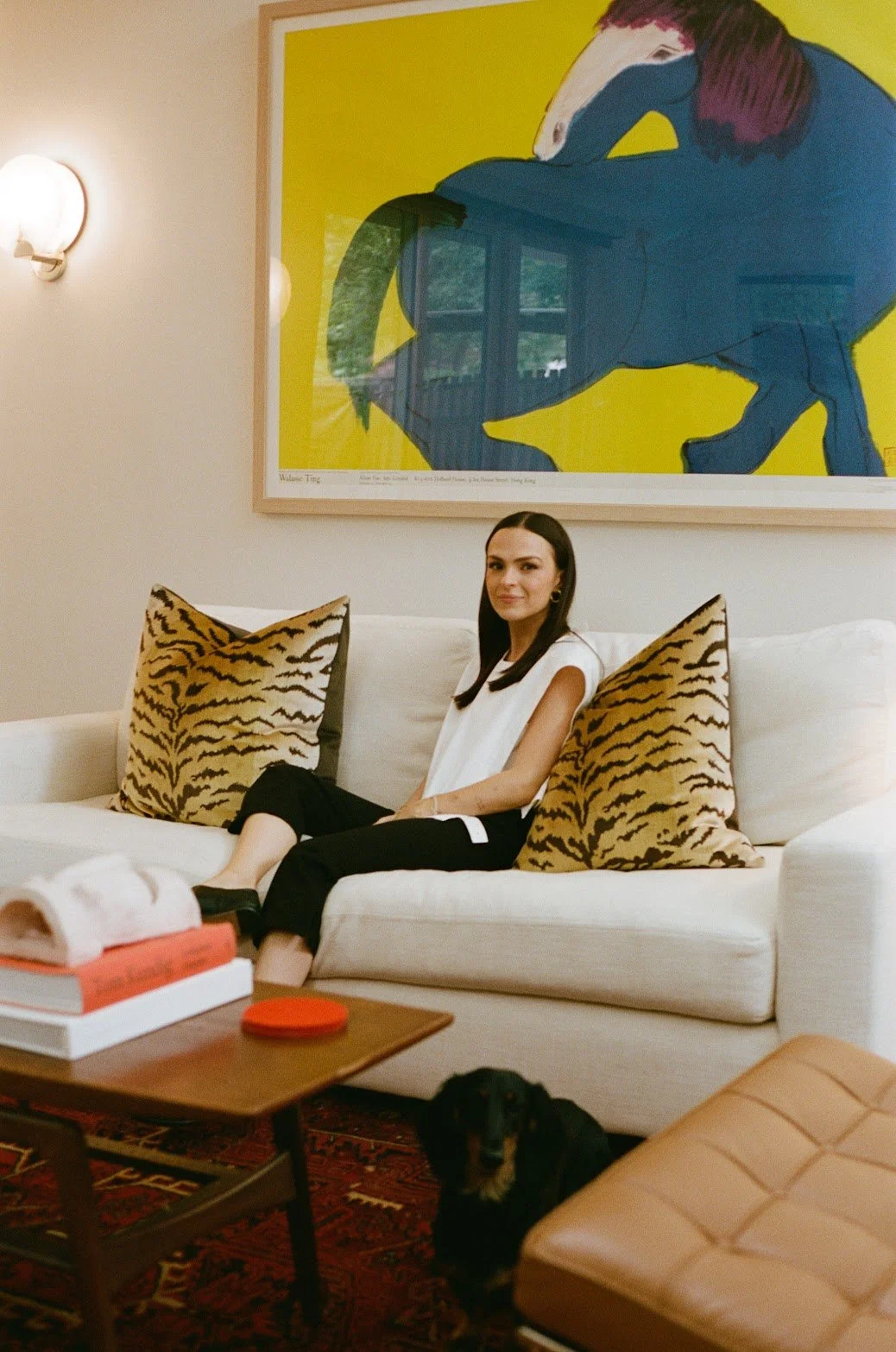 Founder of EMD STUDIO (Emily Mauldin Design Studio) Emily Mauldin, designer and creative, sitting on a white sofa with Scalamandre Le Tigre pillows, a dachshund on a vintage Turkish rug, and a large framed artwork of a horse on the wall behind her.