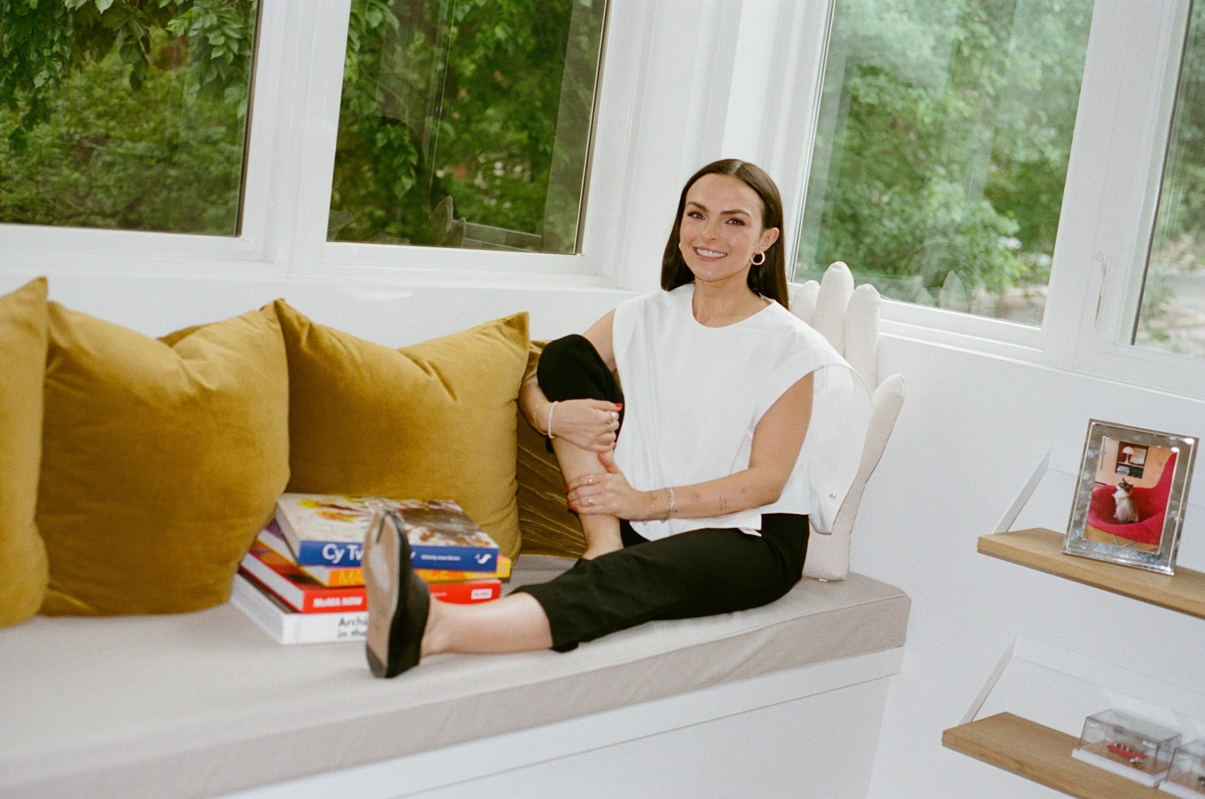 Founder of EMD STUDIO (Emily Mauldin Design Studio) Emily Mauldin, designer and creative, sitting on a window ledge surrounded by yellow cushions, smiling at the camera, with a stack of books and a framed photograph nearby, in a bright room with larg