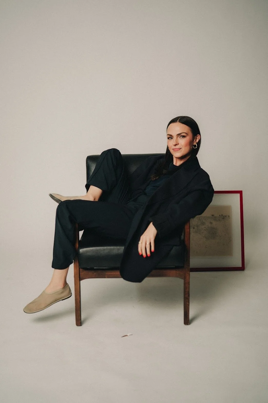 Emily Mauldin, designer and creative, wearing a black blazer and pants, sits on a black leather chair with one leg crossed over the other in a plain room with a framed Raoul Dufy resting on the floor behind her.