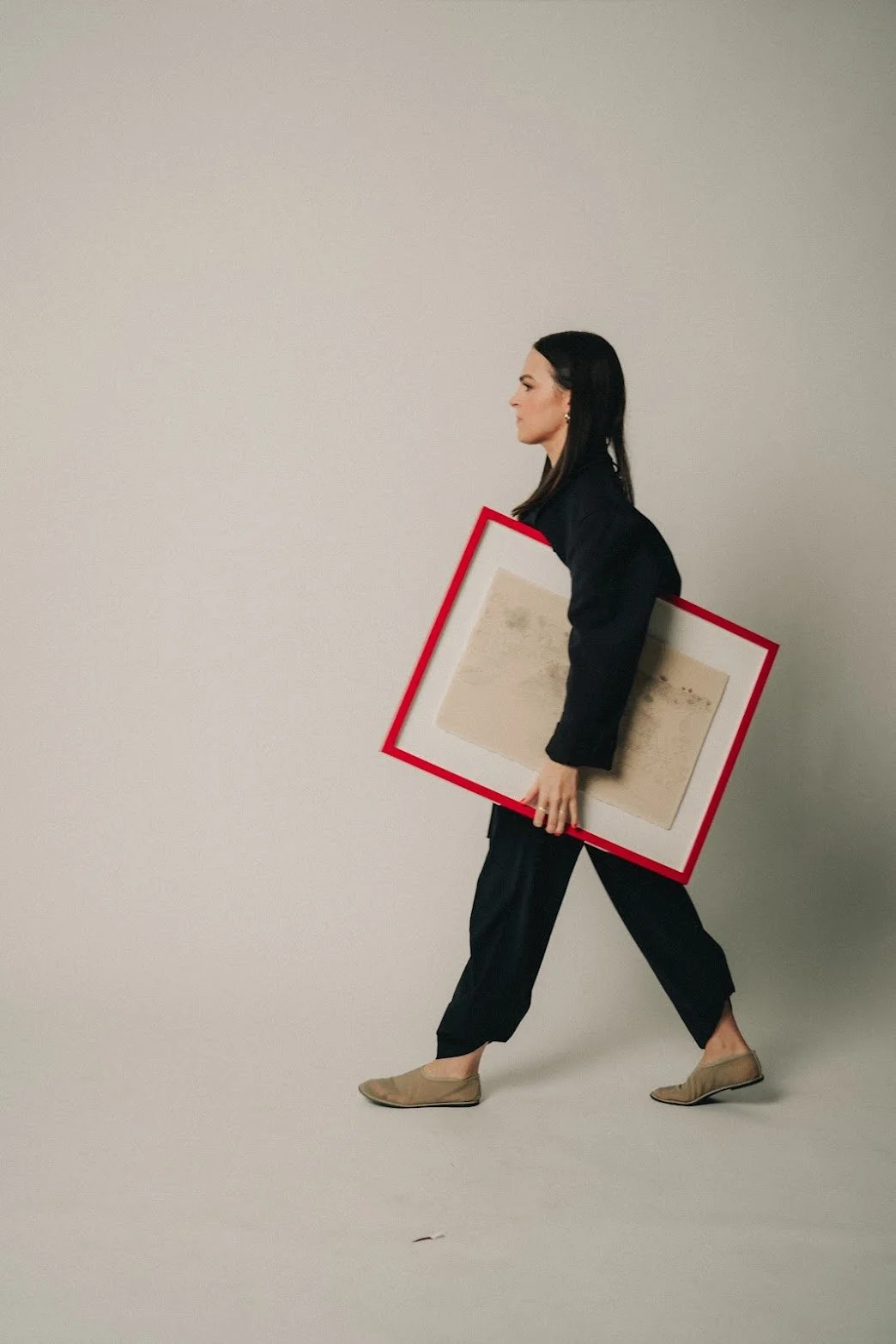 Founder of EMD STUDIO (Emily Mauldin Design Studio), Emily Mauldin, designer and creative, walking sideways carrying an abstract painting with a red border.