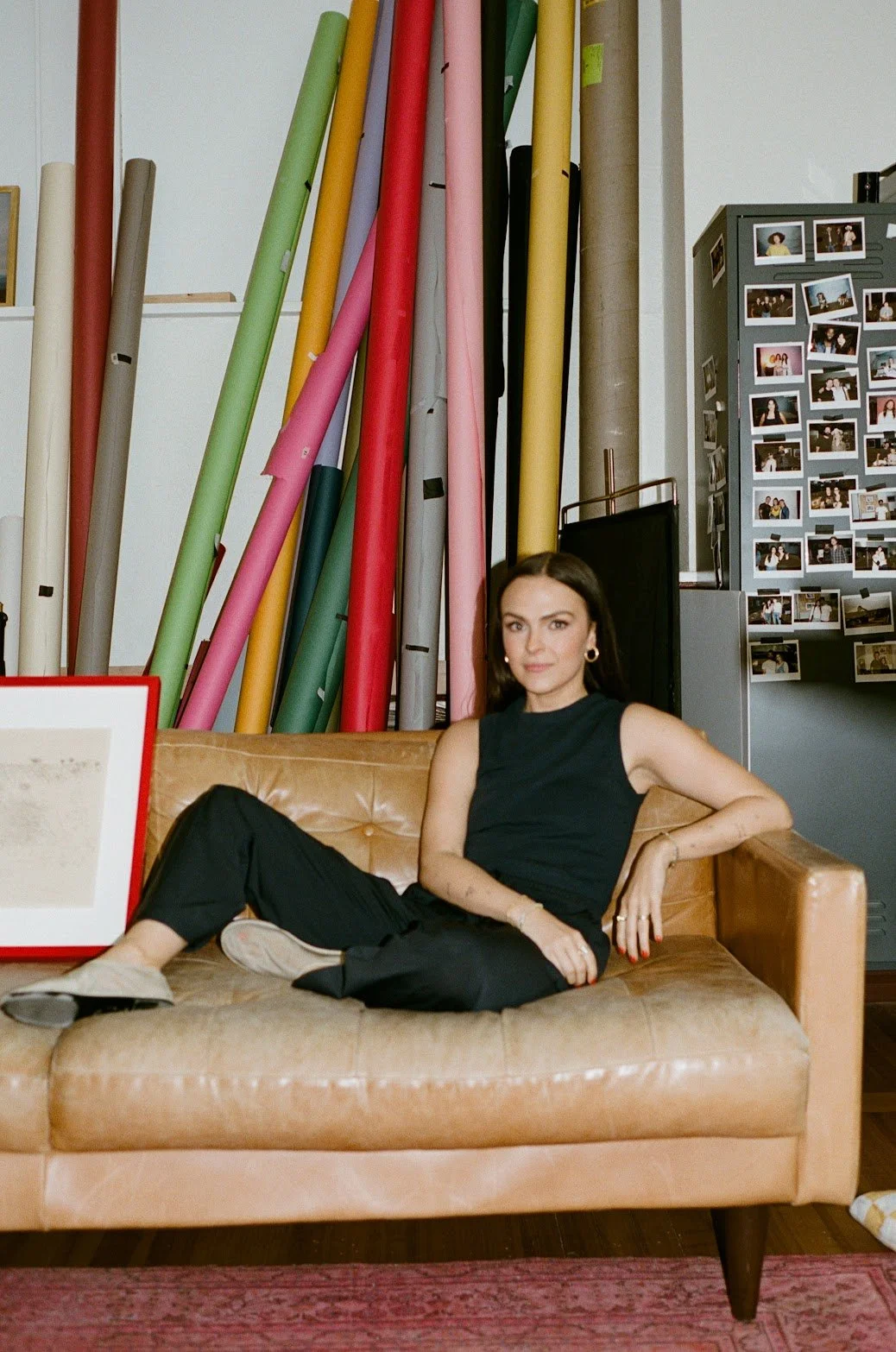 Founder of EMD STUDIO (Emily Mauldin Design Studio), Emily Mauldin, designer and creative, sitting on a tan leather couch in front of large colorful rolls of paper and a wall of photographs.
