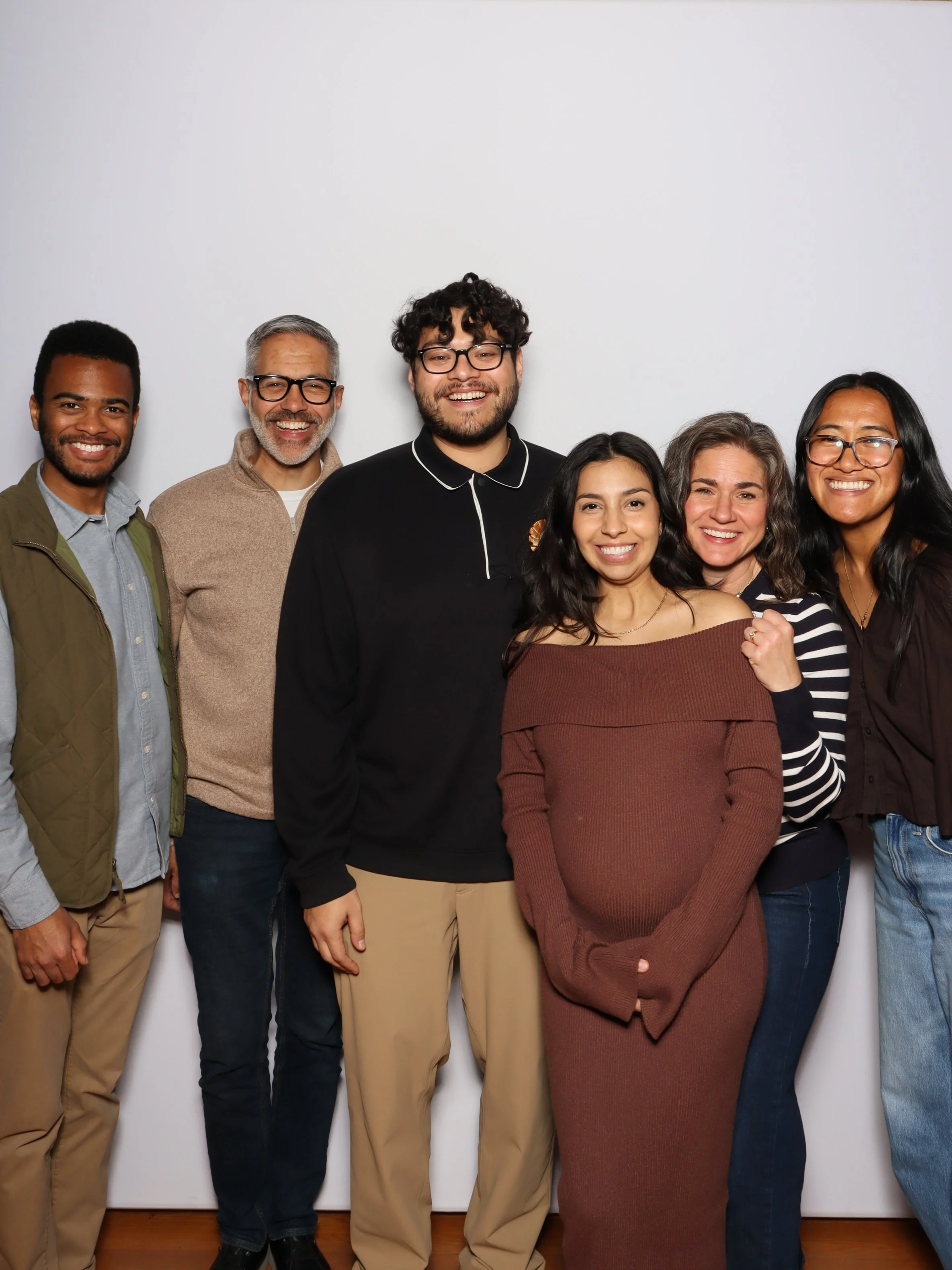 Group of six diverse people smiling and posing together indoors against a plain white wall.