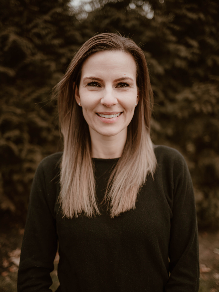 A woman with straight, shoulder-length hair, wearing a black sweater, smiling outdoors in front of a blurred background of trees.