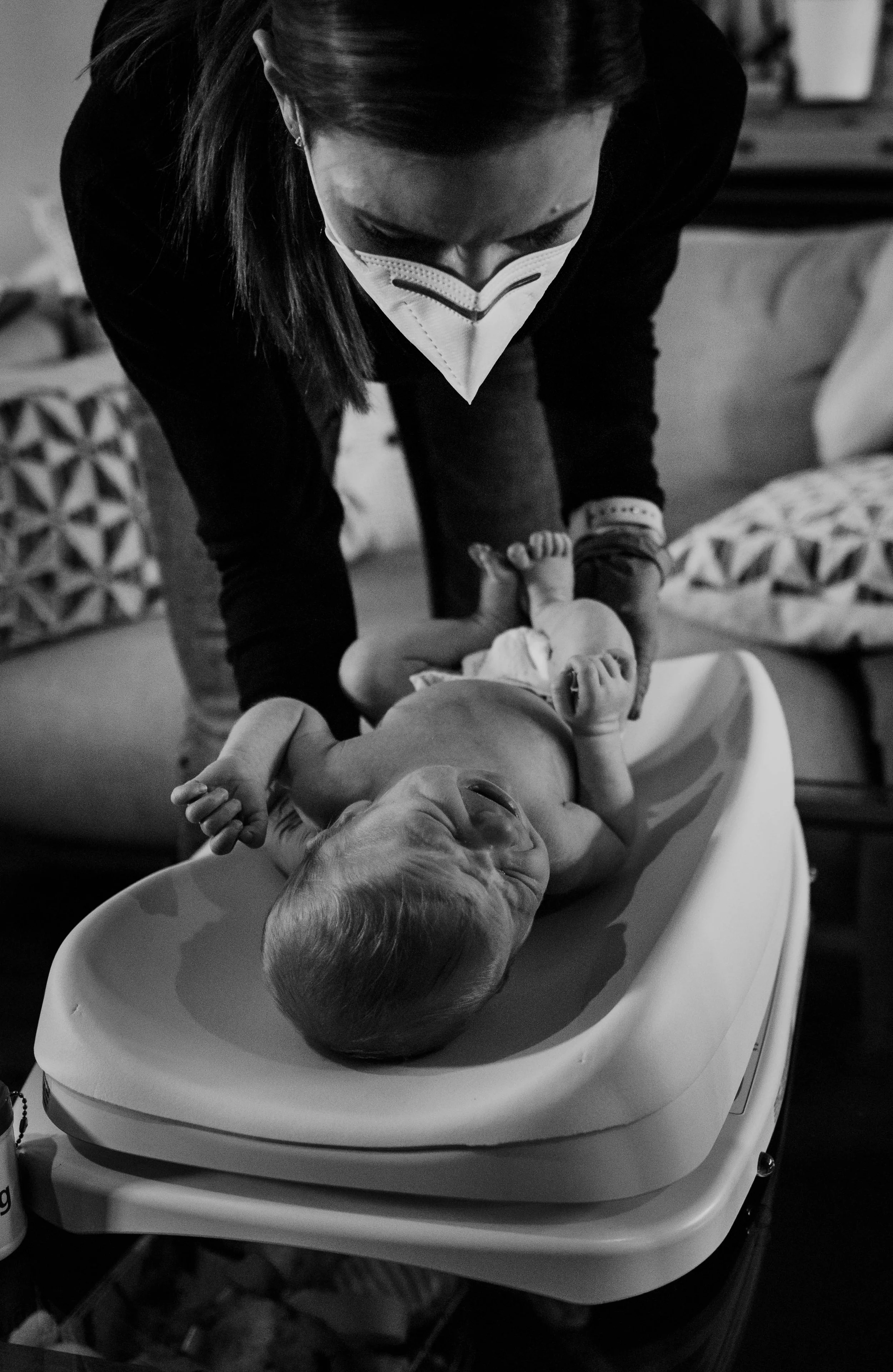 A woman wearing a face mask is leaning over a baby lying on a changing table, laughing or smiling.