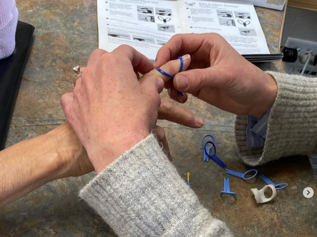 Two people are practicing hand reflexology with hundreds of small, colorful rubber bands on their fingers. A sheet with instructions and diagrams is on the table in the background.