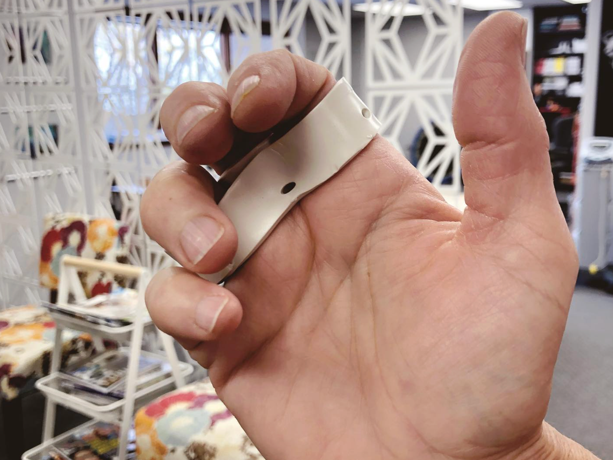 Close-up of a person's hand holding a small, silver, circular object with holes, possibly a vintage camera lens cap, indoor setting with decorative white lattice panels and shelves in the background.