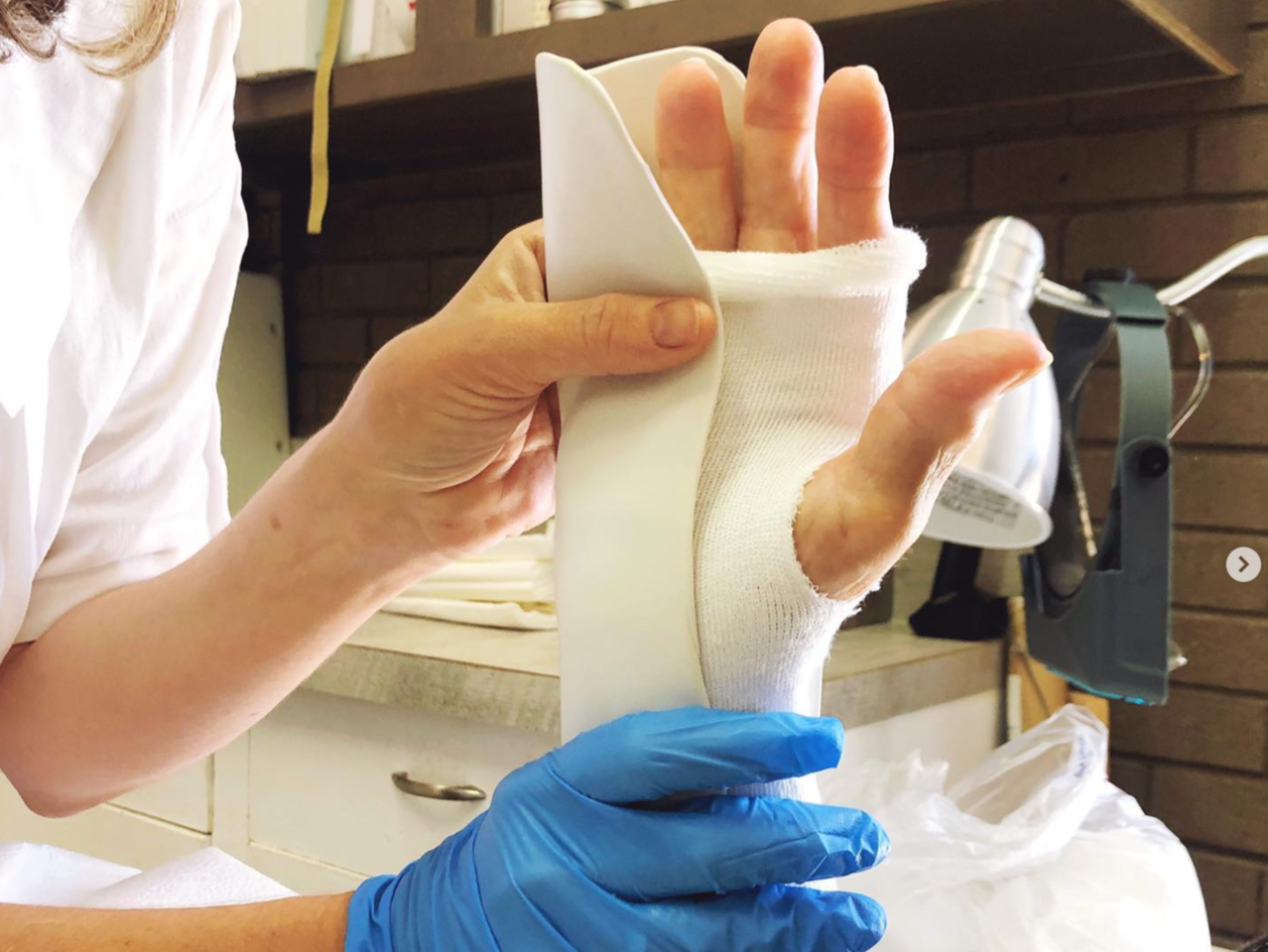 A person is wrapping a bandage around a person's injured hand, with another person assisting while wearing blue gloves. The background shows a brick wall and medical equipment.