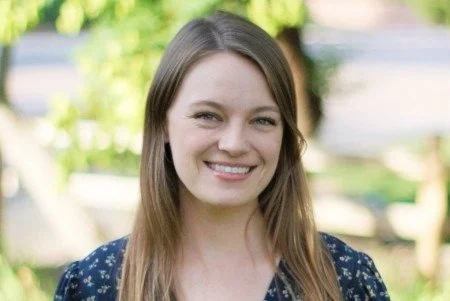 A young woman with long brown hair smiling outdoors with greenery and trees in the background.