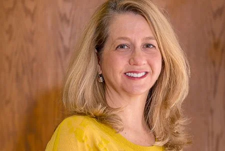 A woman with blonde hair wearing a yellow top, smiling, in front of a wooden background.