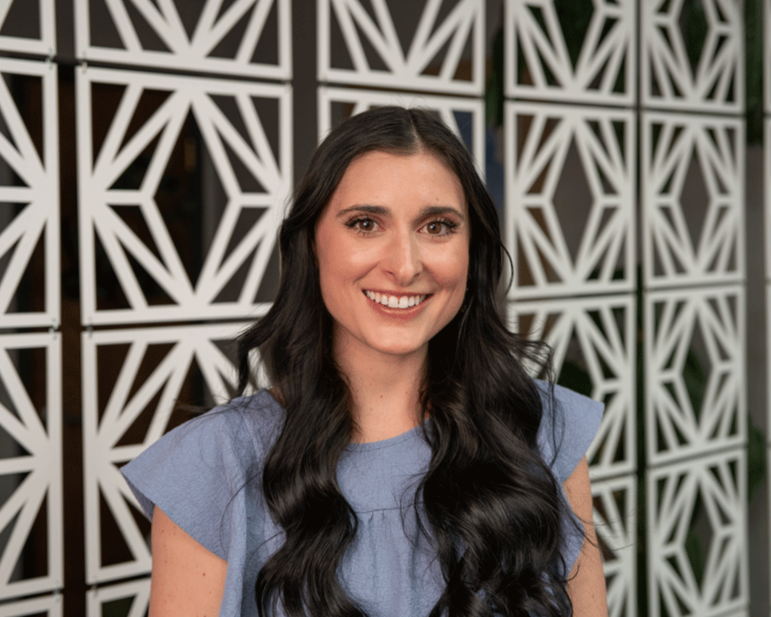 A woman with long dark hair smiling at the camera, standing against a light-colored brick wall