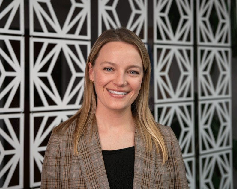 A young woman with long brown hair smiling outdoors with greenery and trees in the background.