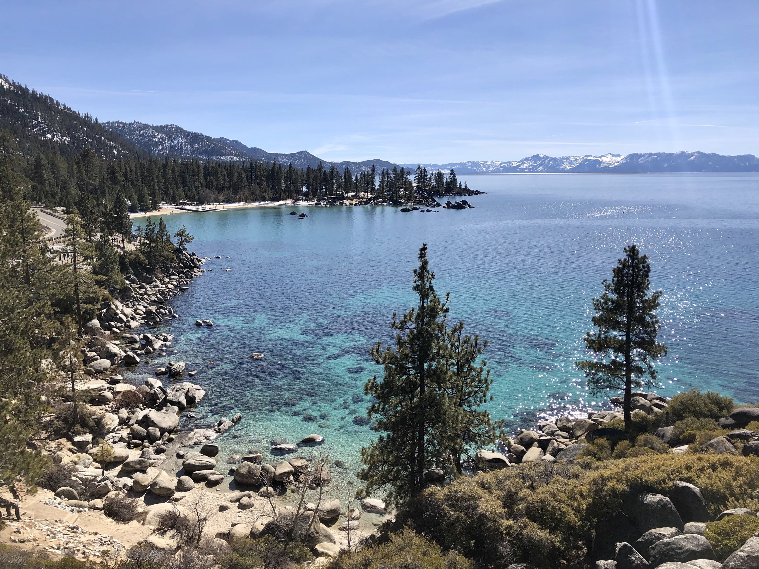 Scenic view of a lake surrounded by mountains and pine trees, with clear blue water and snow-capped peaks in the distance.