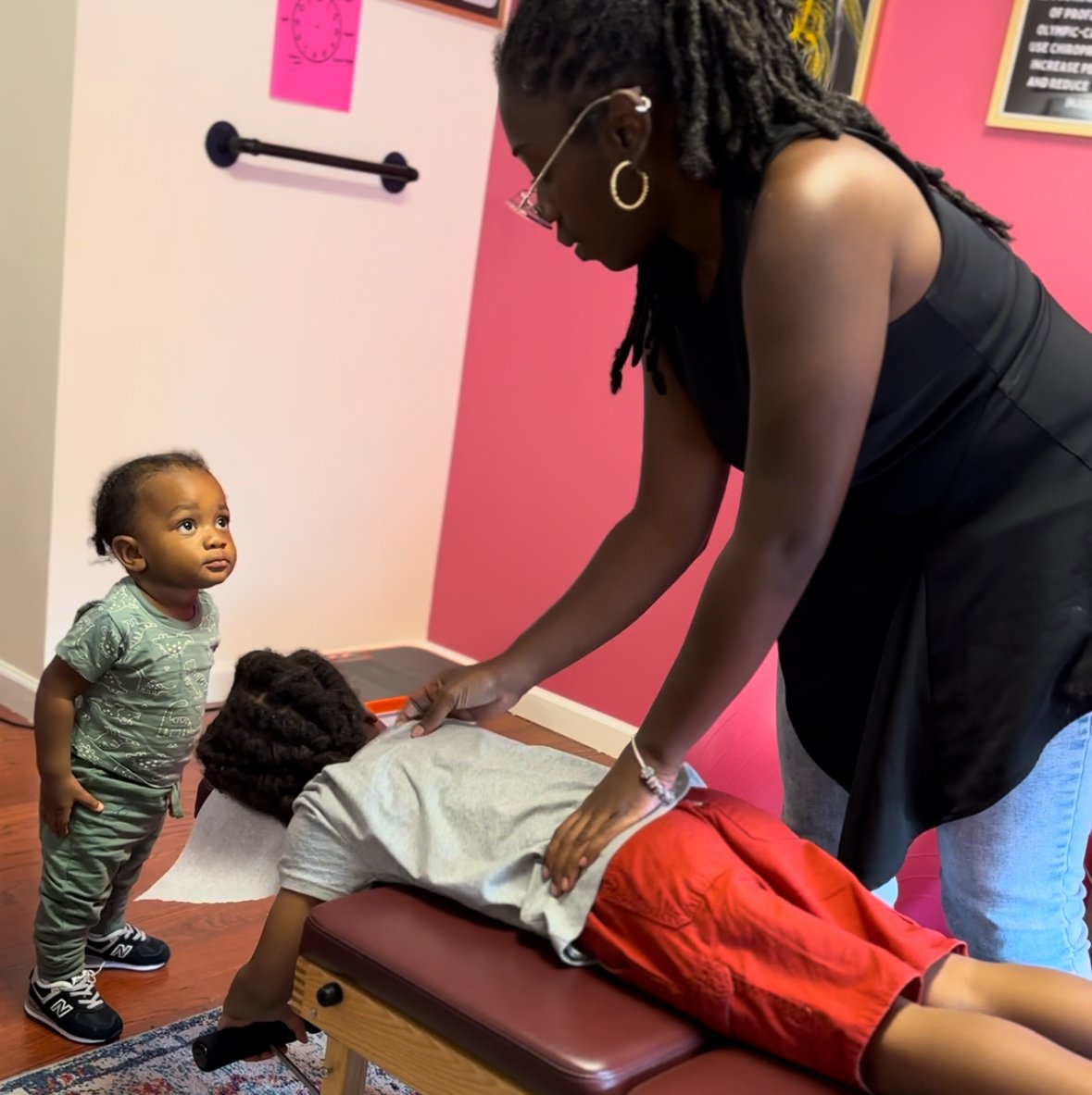 Dr. Magen providing a pediatric chiropractic adjustment to a child lying face down on a chirorpactic table while a young boy stands nearby in a room with pink and white walls.
