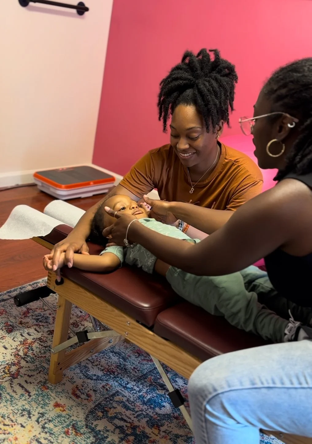 Dr. Magen Henry, examining a young boy lying on a chiropractic table in a room with pink walls, with one woman smiling and the other touching the boy's face, suggesting a pediatric chiropractic therapy.