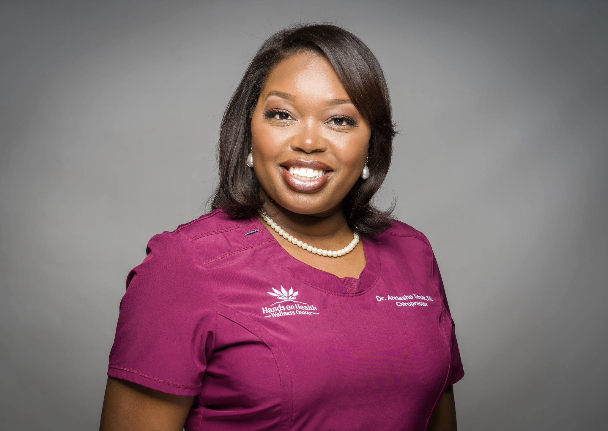 A woman with medium-length dark hair smiling, wearing pearl earrings, a pearl necklace, and a magenta top with embroidered text that reads 'Hands On Health Wellness Center' and 'Dr. Amiesha Scott, DC, Chiropractor' set against a gray background.