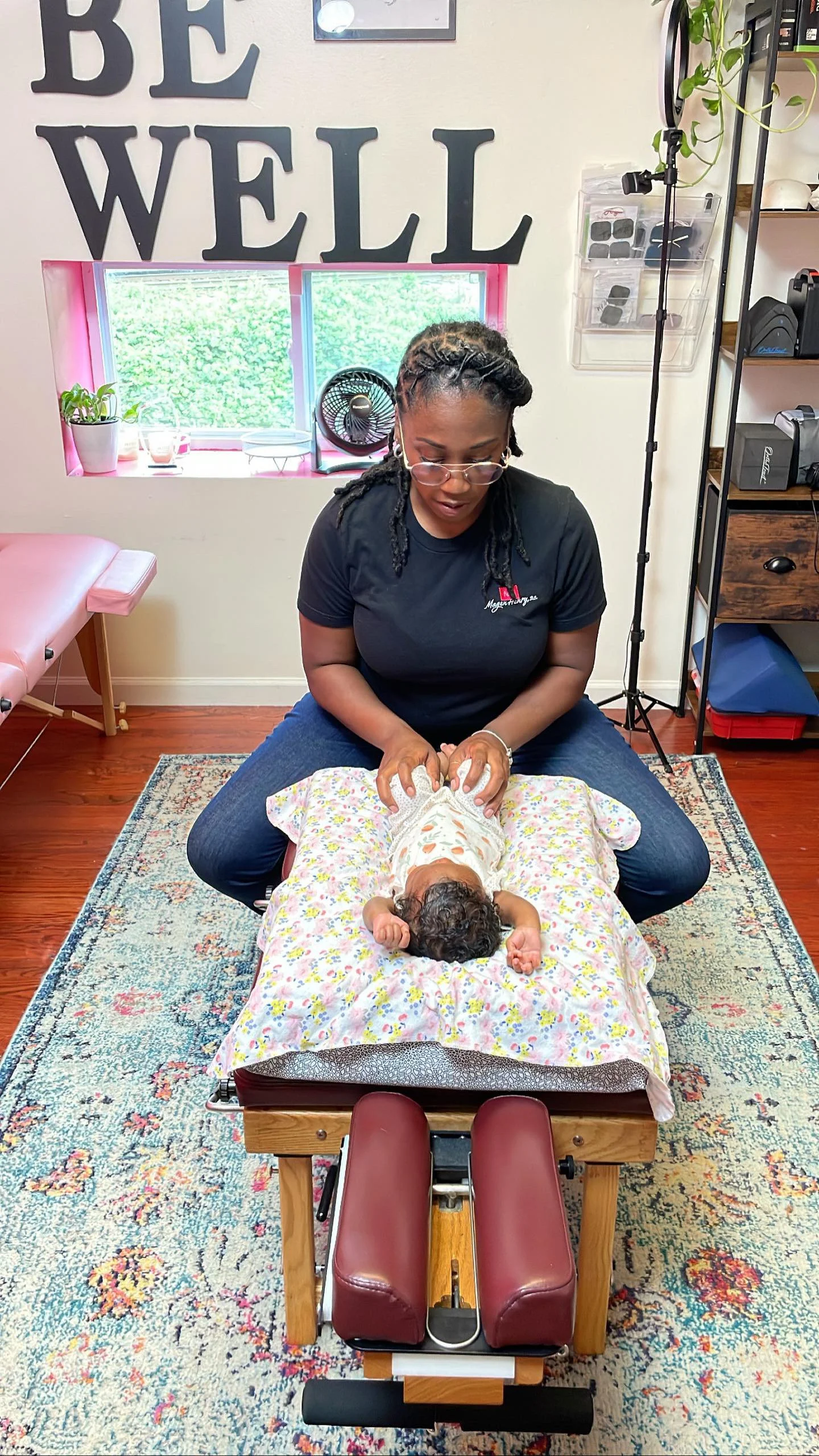 A woman practicing chiropractic adjustments on a baby lying face down on a padded table in a wellness chiropractic clinic. The child is covered with a colorful blanket, and the woman is wearing glasses and a black T-shirt. The clinic has a sign that 