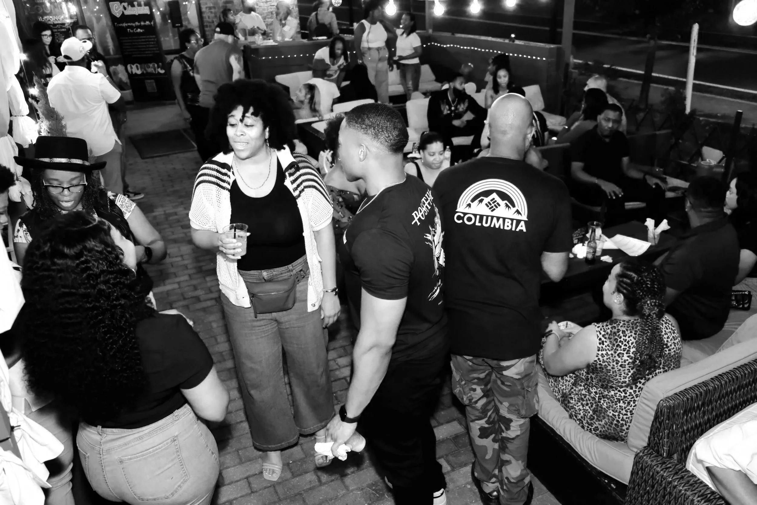 A group of people socializing outdoors at night at a bar or restaurant. Some are standing, some are seated, with a bar visible in the background and string lights overhead.