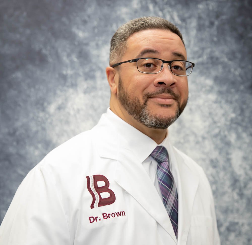 Professional man with glasses, beard, and mustache, wearing a white lab coat with 'Dr. Brown' embroidered on it, a striped tie, and standing against a neutral background.