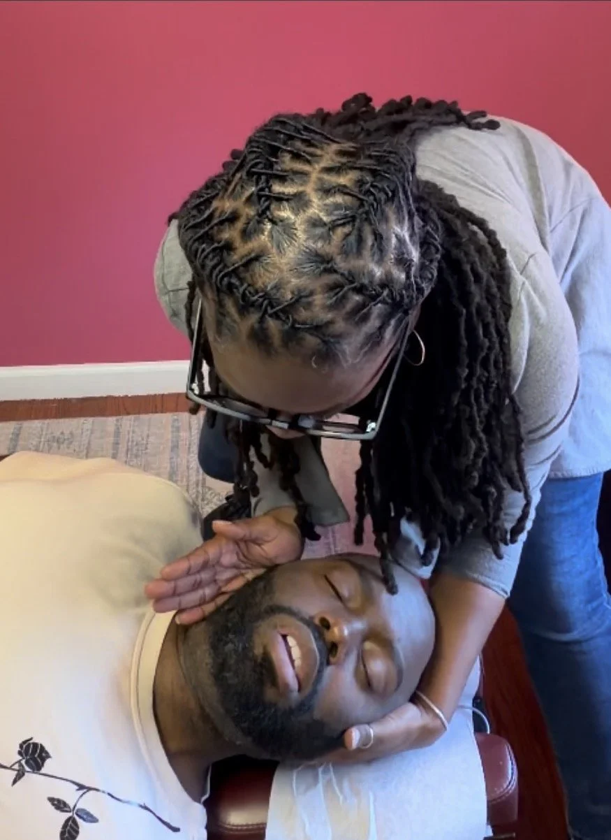 Woman with dreadlocks giving a gentle massage or attend to a man who is lying down on a massage table with eyes closed, appearing relaxed, against a pink wall.