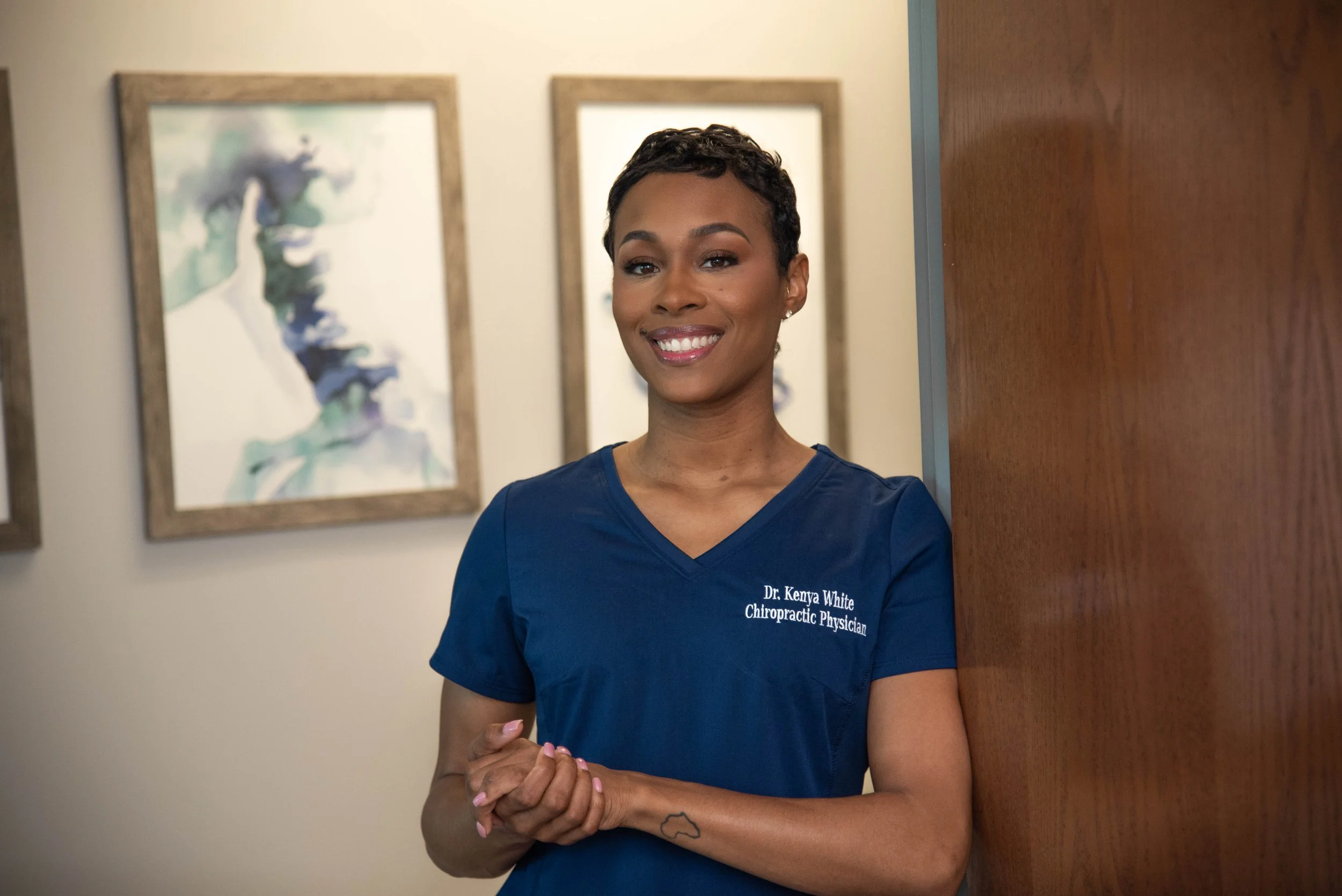 A smiling African American woman in blue medical scrubs standing in a hallway near a wooden wall, with framed abstract artwork behind her.