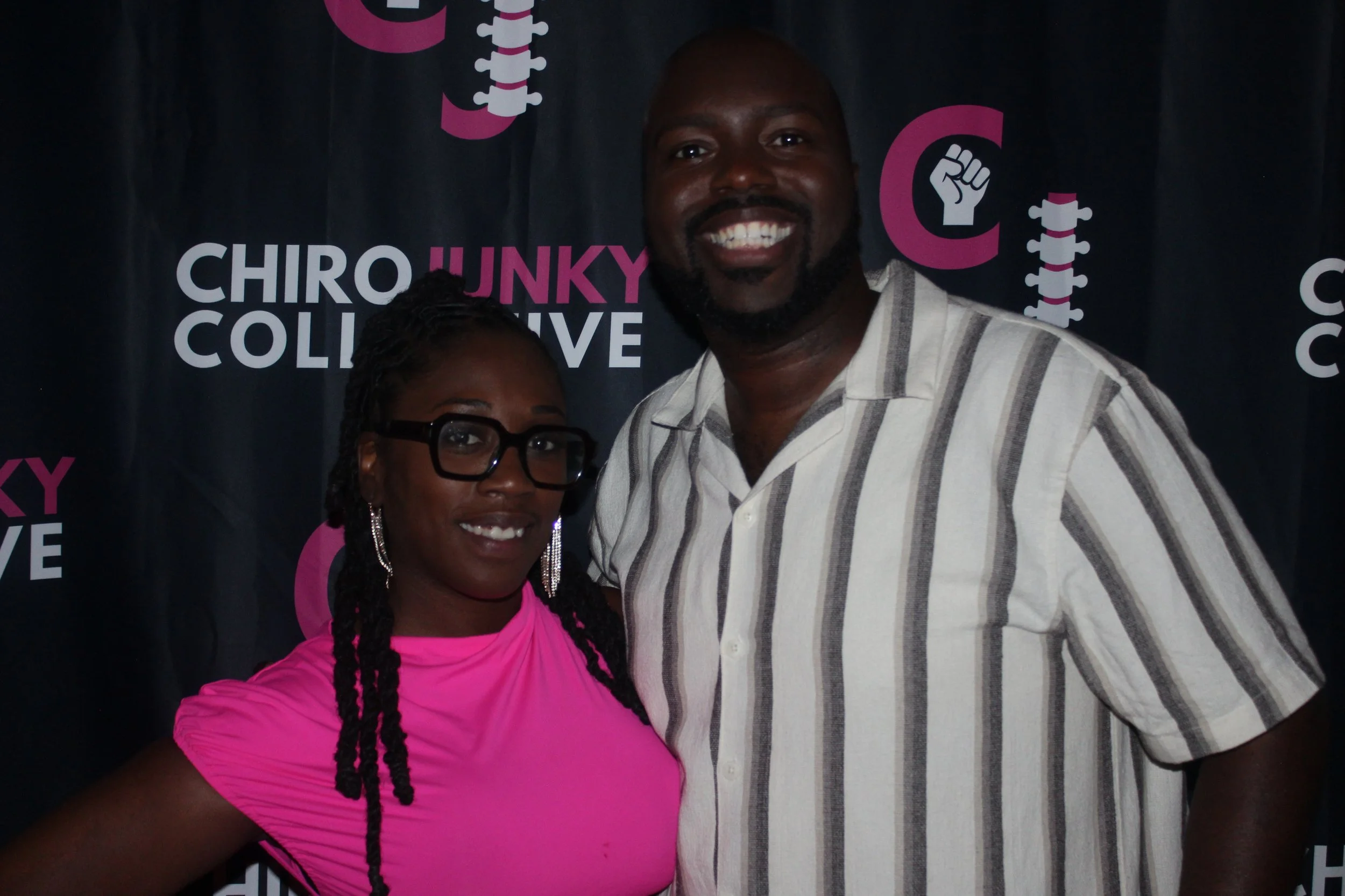 Two people smiling and posing for a photo in front of a dark backdrop with text and graphics. The woman on the left wears glasses, large earrings, and a bright pink top. The man on the right wears a striped short-sleeve shirt.