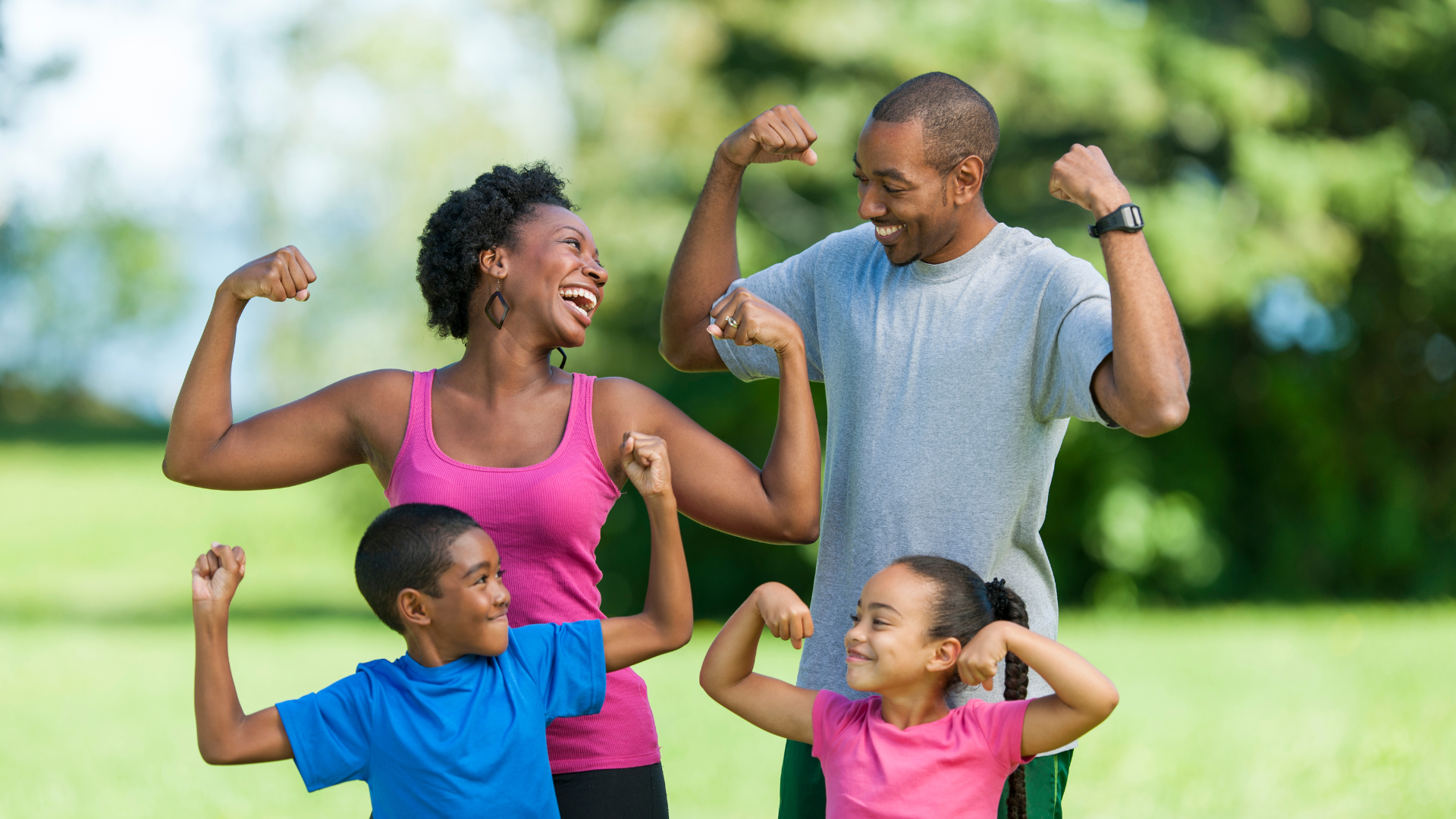 A happy family of four outdoors, flexing their muscles and smiling at each other, with a background of green trees and grass.