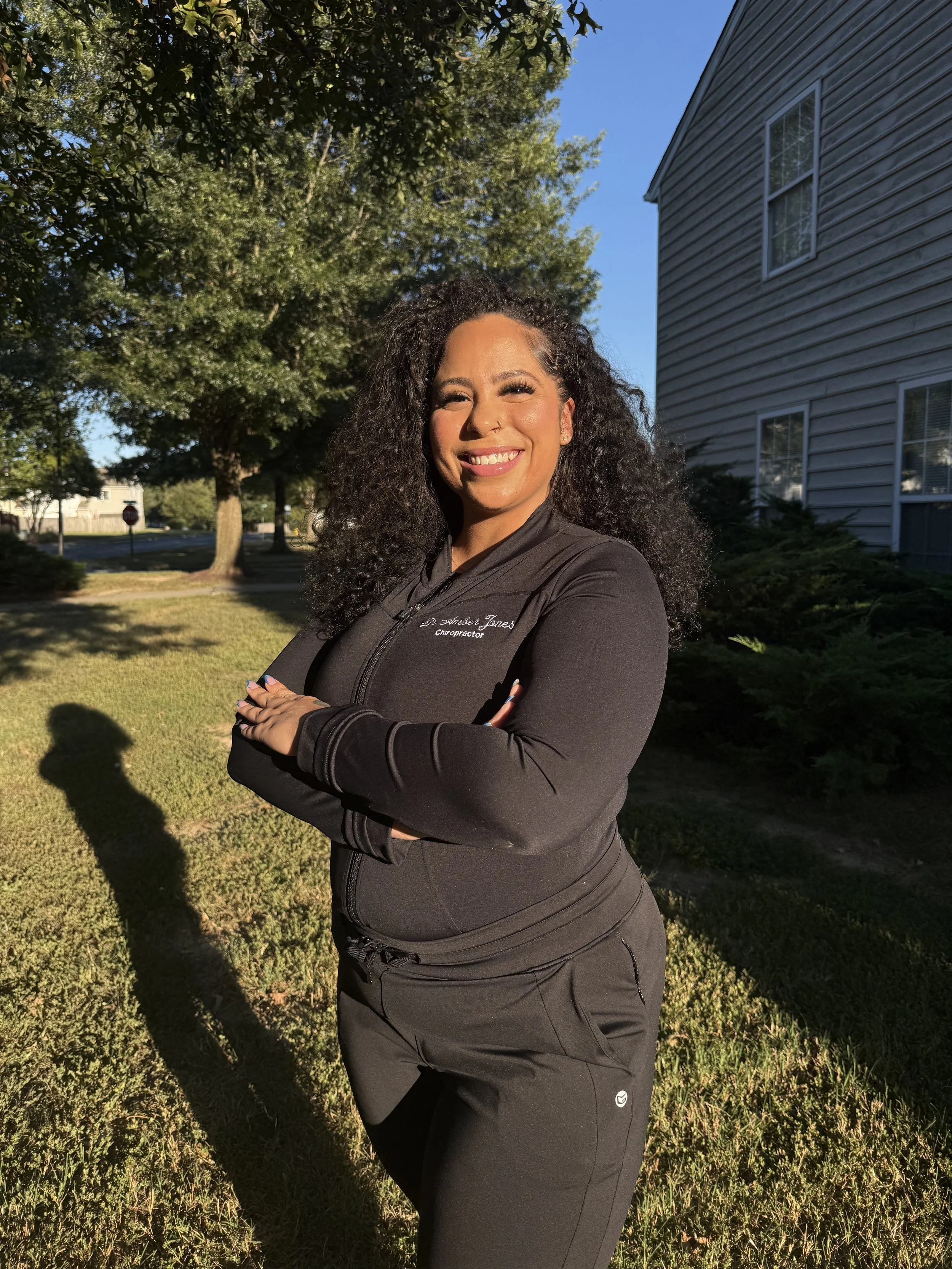 A woman with curly hair smiling outdoors in the late afternoon sunlight, wearing a black zip-up jacket and black pants, standing with arms crossed in front of a house and green trees.