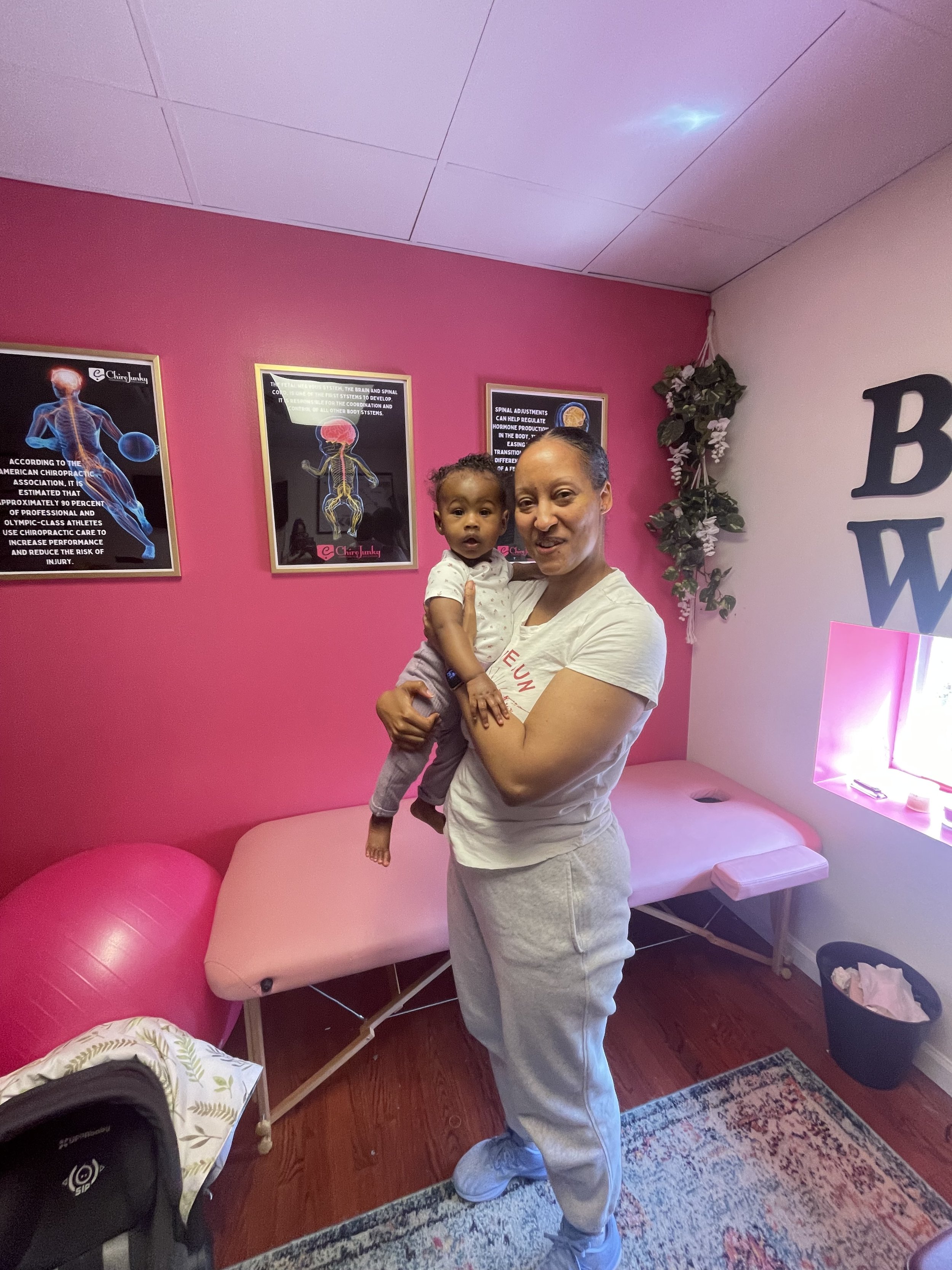 A woman holding a young child in a room with pink and white walls, framed posters about chiropractic care, a pink therapy table, an exercise ball, and a rug on wooden floor.