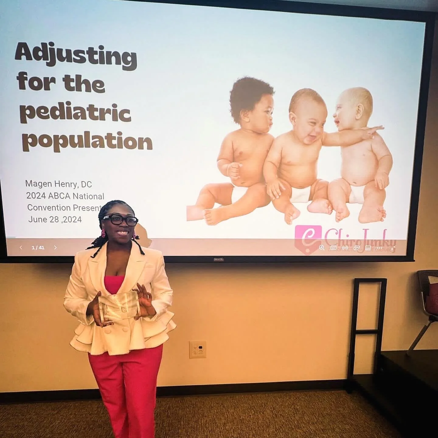 Dr. Magen giving a presentation in front of a large screen displaying a slide titled 'Adjusting for the pediatric population.' The slide includes an image of three babies sitting and interacting with each other.