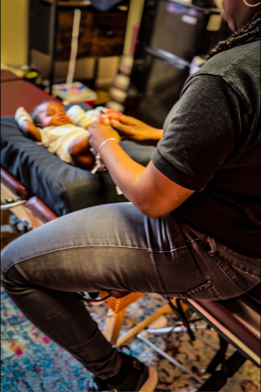Dr. Magen Henry sitting on a stool checking a baby's feet while the baby lies on a chiropractic table in a clinic.