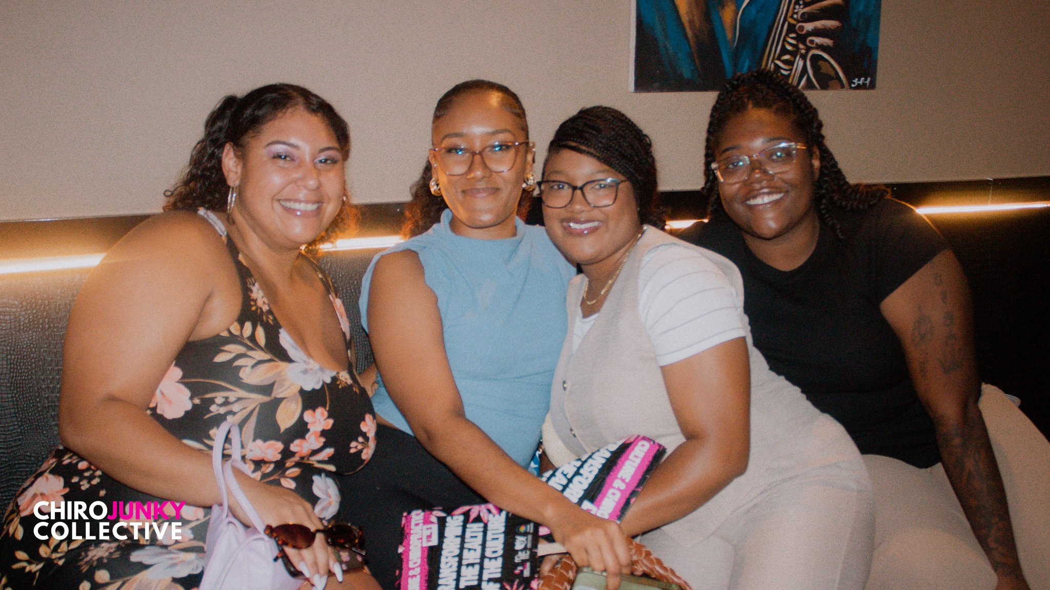 Four women sitting together on a black bench, smiling for the camera in a dimly lit room, with a colorful abstract painting on the wall behind them.