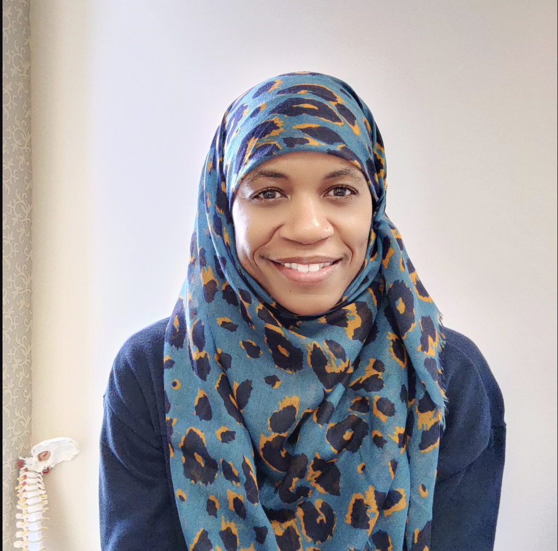 A woman with a blue leopard print hijab and dark clothing smiling in front of a plain white wall, with a spine model partially visible on the left side.