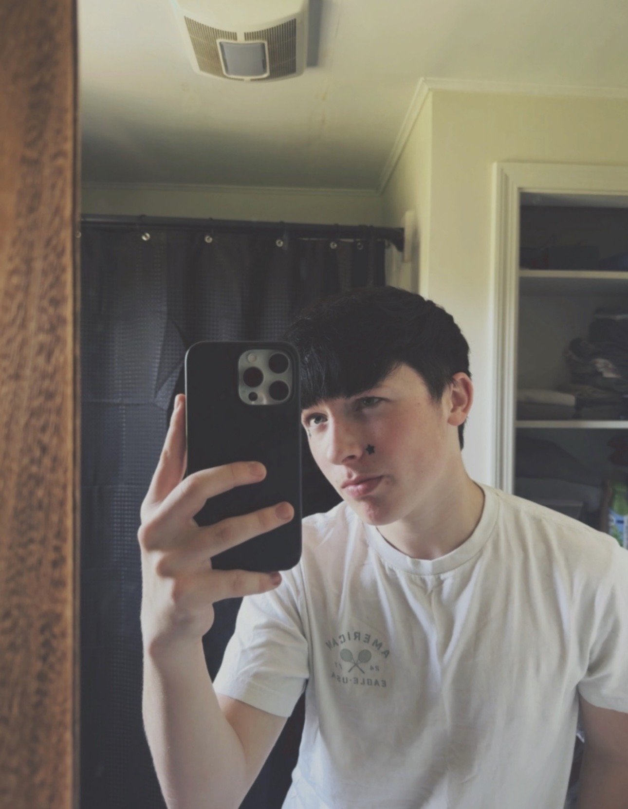 Young man taking a selfie in a bathroom mirror, holding a smartphone in front of his face and wearing a white T-shirt.