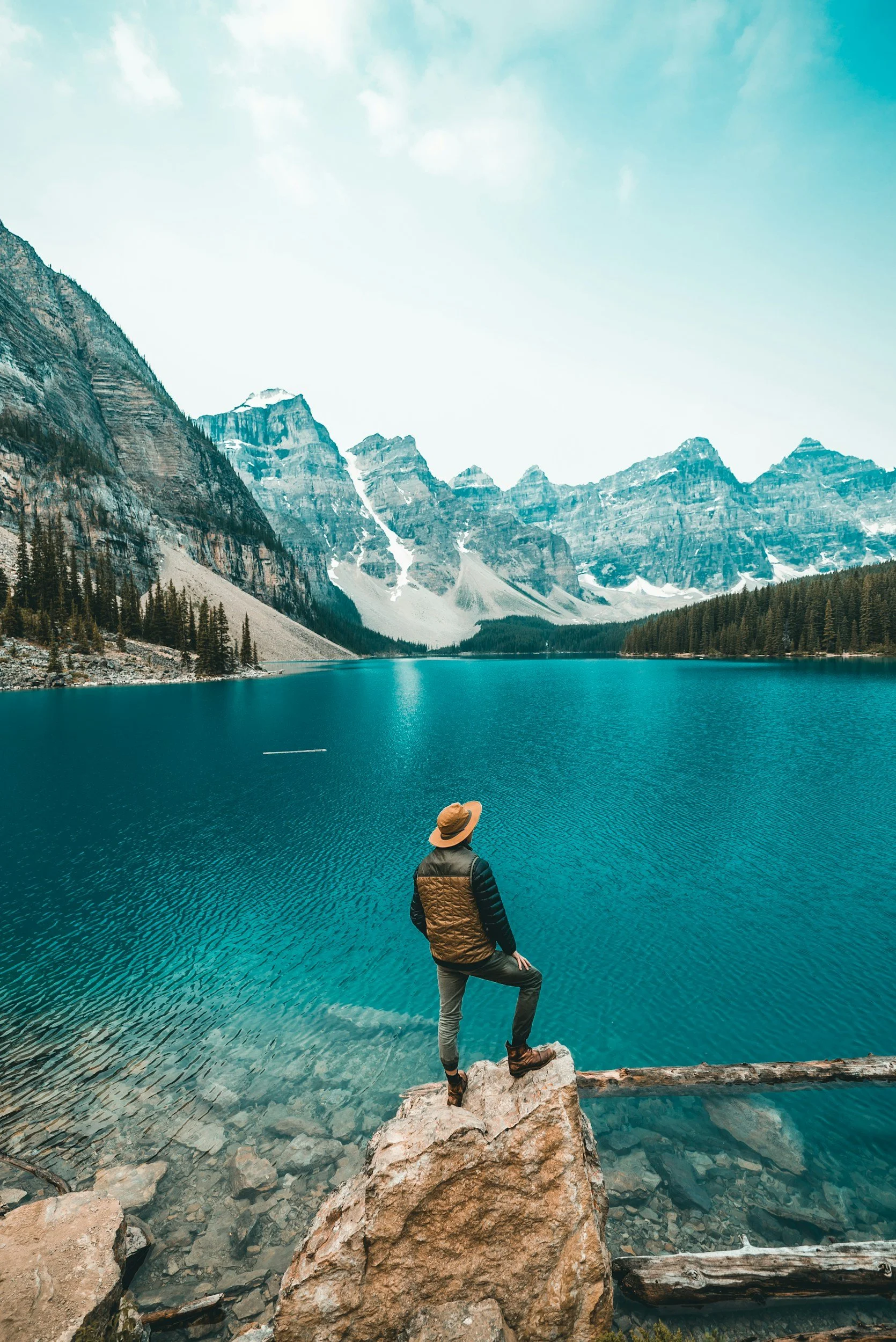 A person standing on rocks by a large, blue mountain lake with mountain peaks in the background and a bright sky overhead.