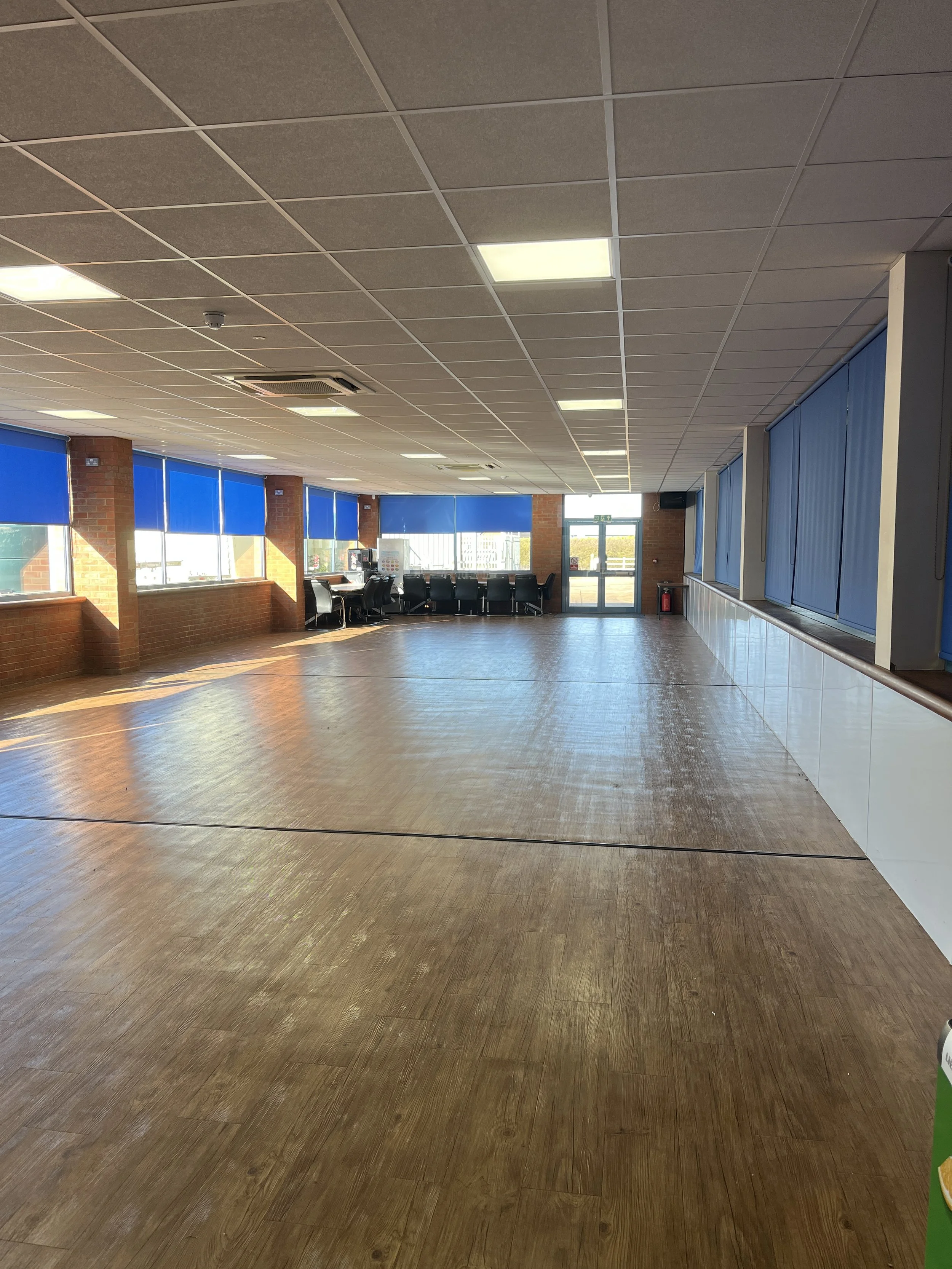 Empty room with wooden flooring, blue window blinds, brick walls, large windows, and a row of chairs near the back wall.