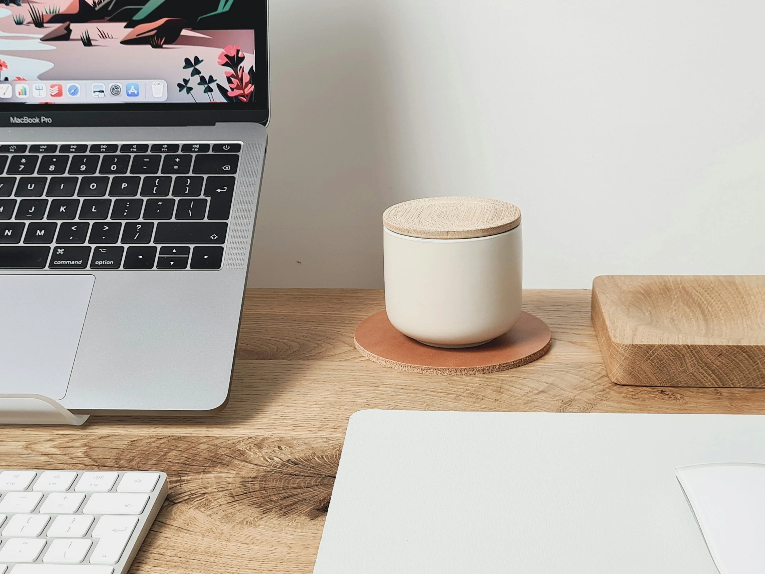 A tidy workspace with a MacBook Pro laptop, a white keyboard, a wooden tray, a ceramic container with a wooden lid on a round leather coaster, and a wooden tray on a light wooden desk.