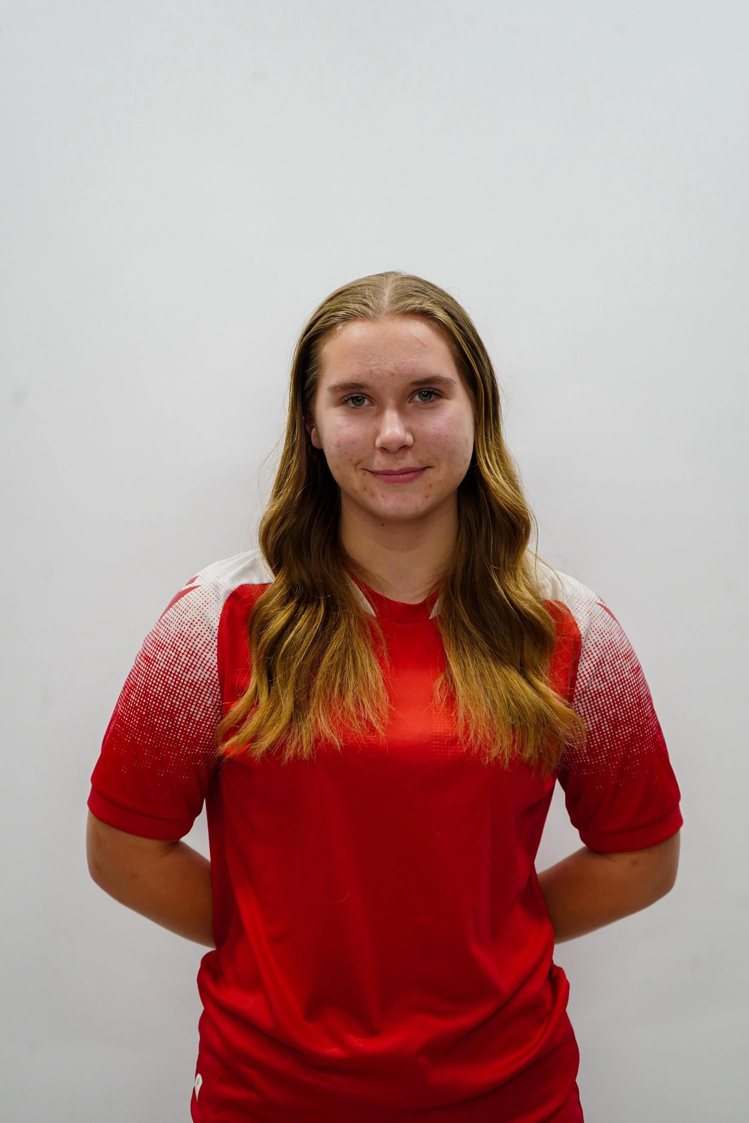 Young woman with long wavy hair wearing a red sports jersey standing in front of a plain white wall.