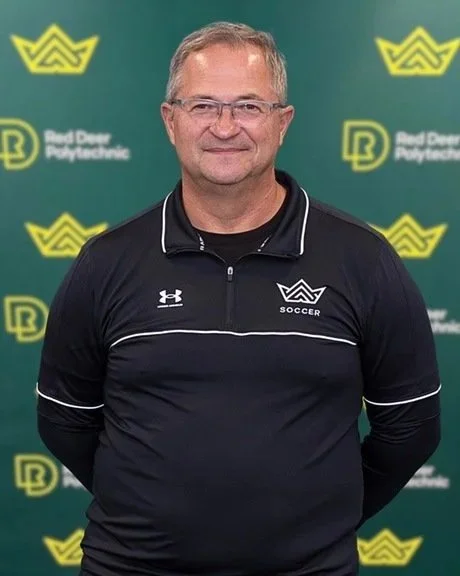 A middle-aged man with glasses, wearing a black Under Armour shirt with white accents, stands in front of a green background with yellow logos and the text "Red Deer Polytechnic" and a soccer emblem.
