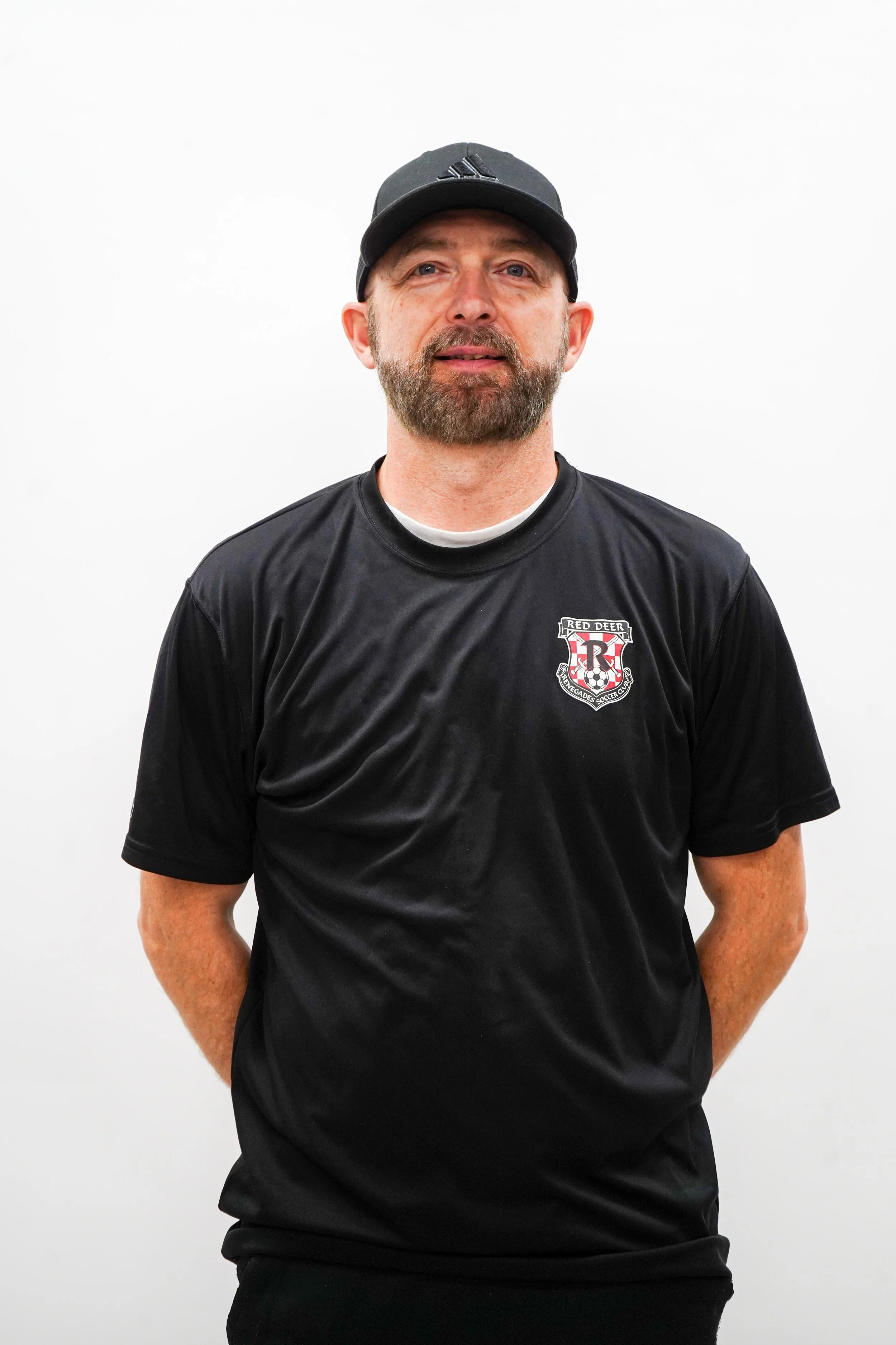 A man with a beard and mustache wearing a black baseball cap and black T-shirt with a red and white logo, standing against a plain white background.