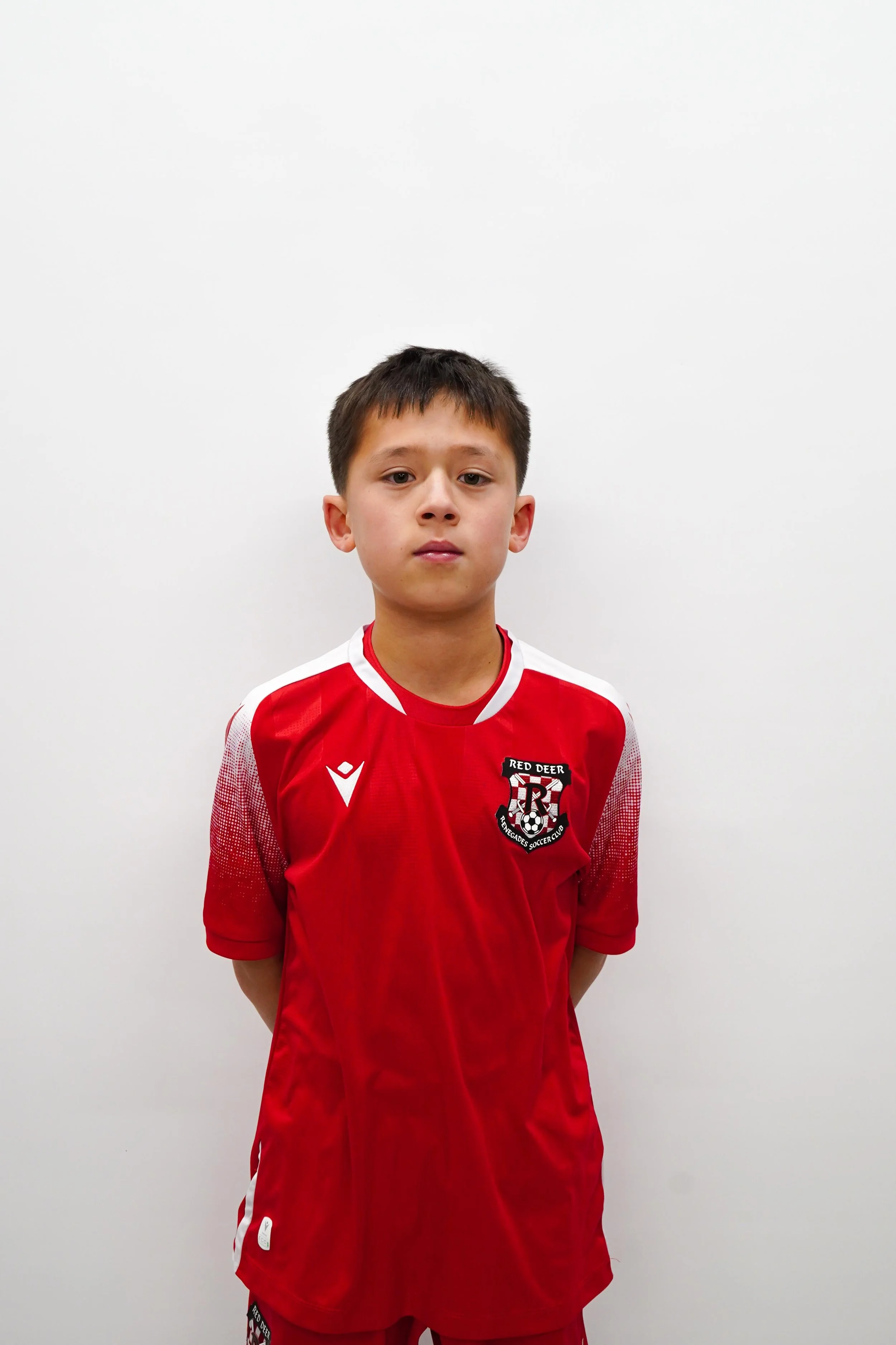 Young boy wearing a red soccer jersey with a 'Red Deer' logo standing against a plain white wall.