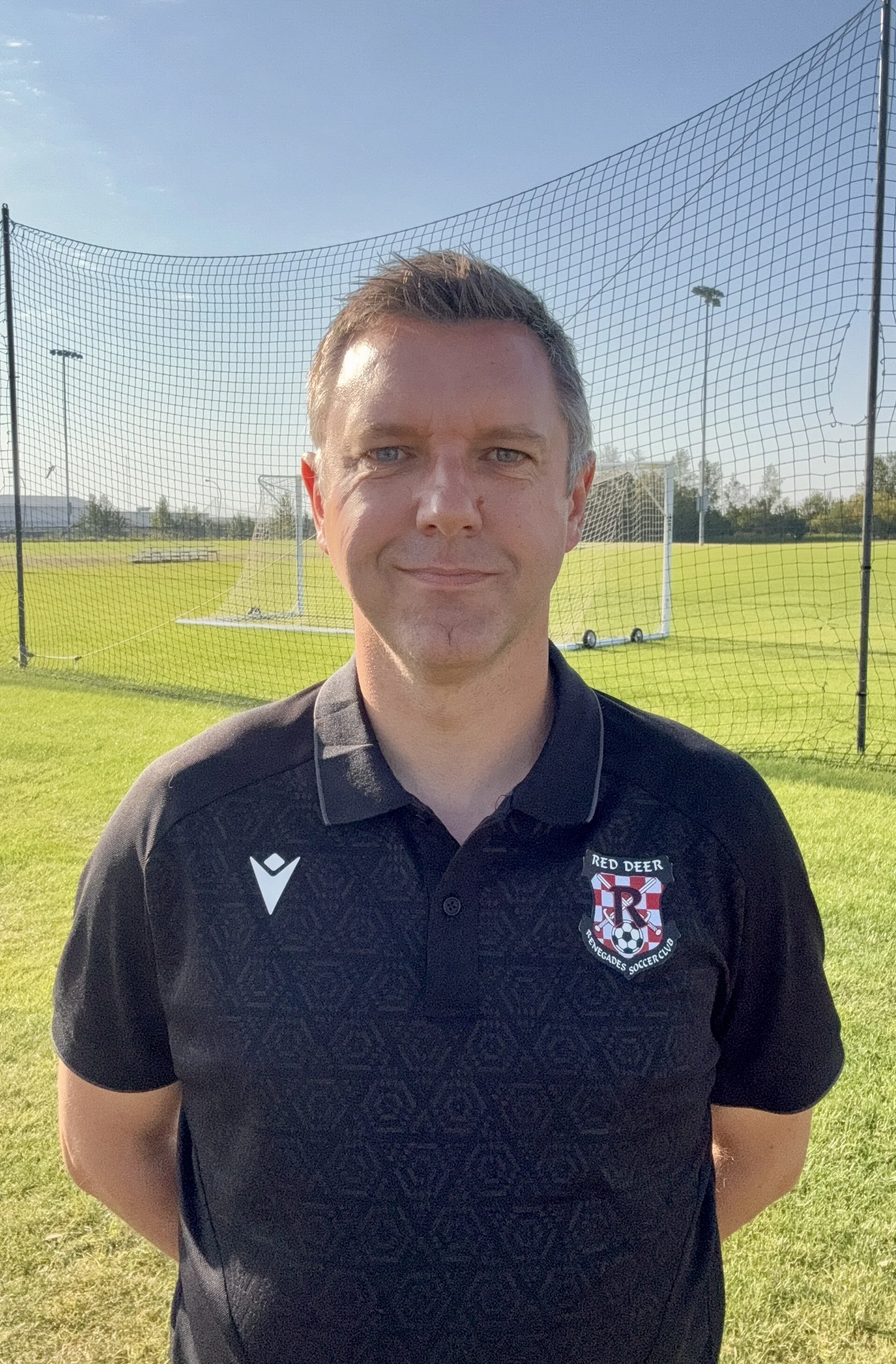 A man standing on a soccer field with goalposts and netting in the background, wearing a black sports polo shirt with a soccer team's emblem.