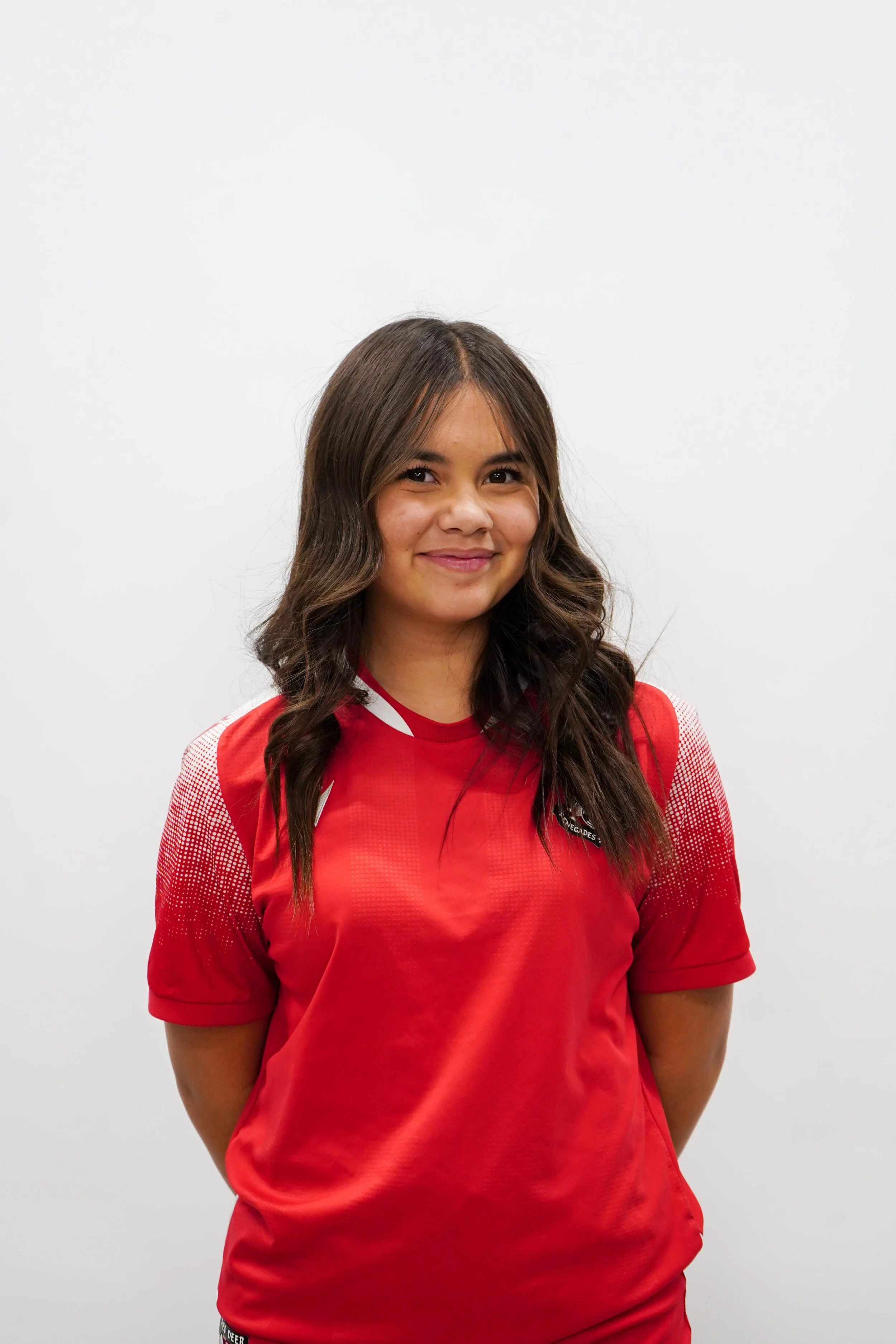Young woman with long brown wavy hair smiling, wearing a red sports jersey, standing against a plain white background.