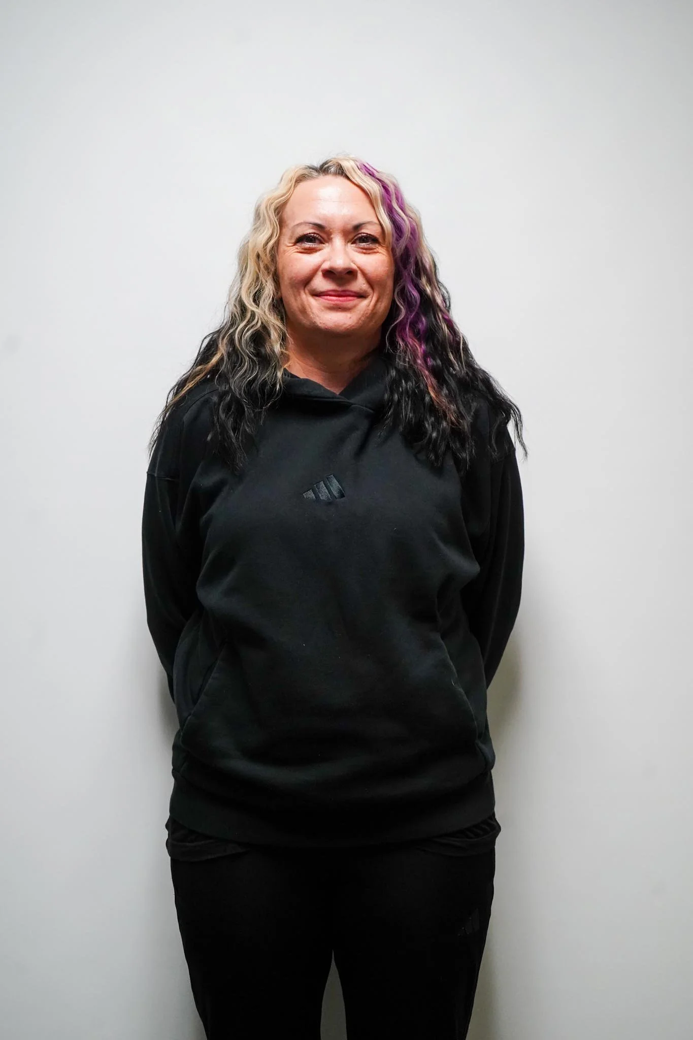 A woman with blonde curly hair streaked with purple and black, wearing a black Adidas hoodie, standing against a plain white wall, smiling slightly.