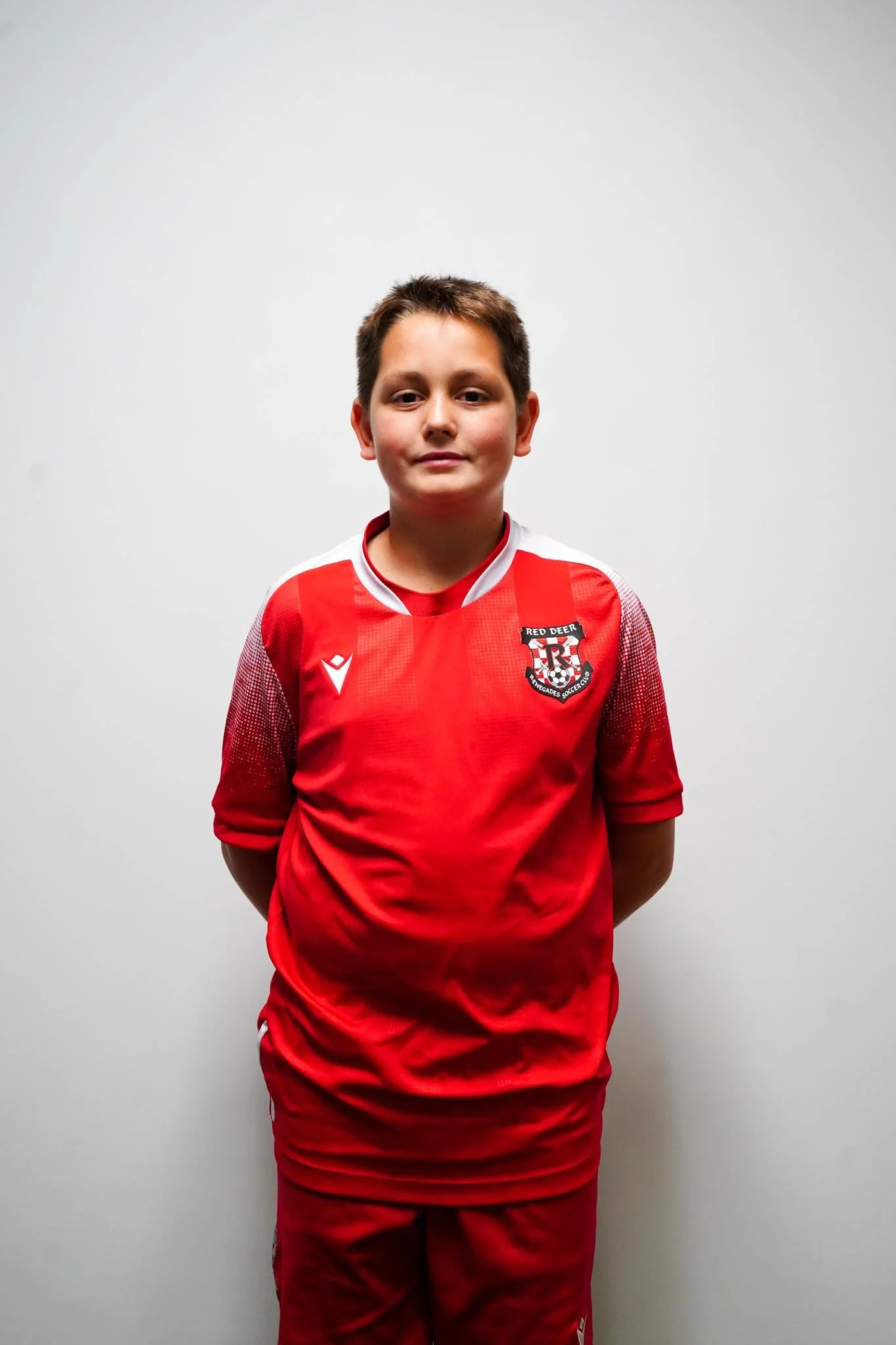 A young boy wearing a red sports jersey and shorts standing against a plain white wall.