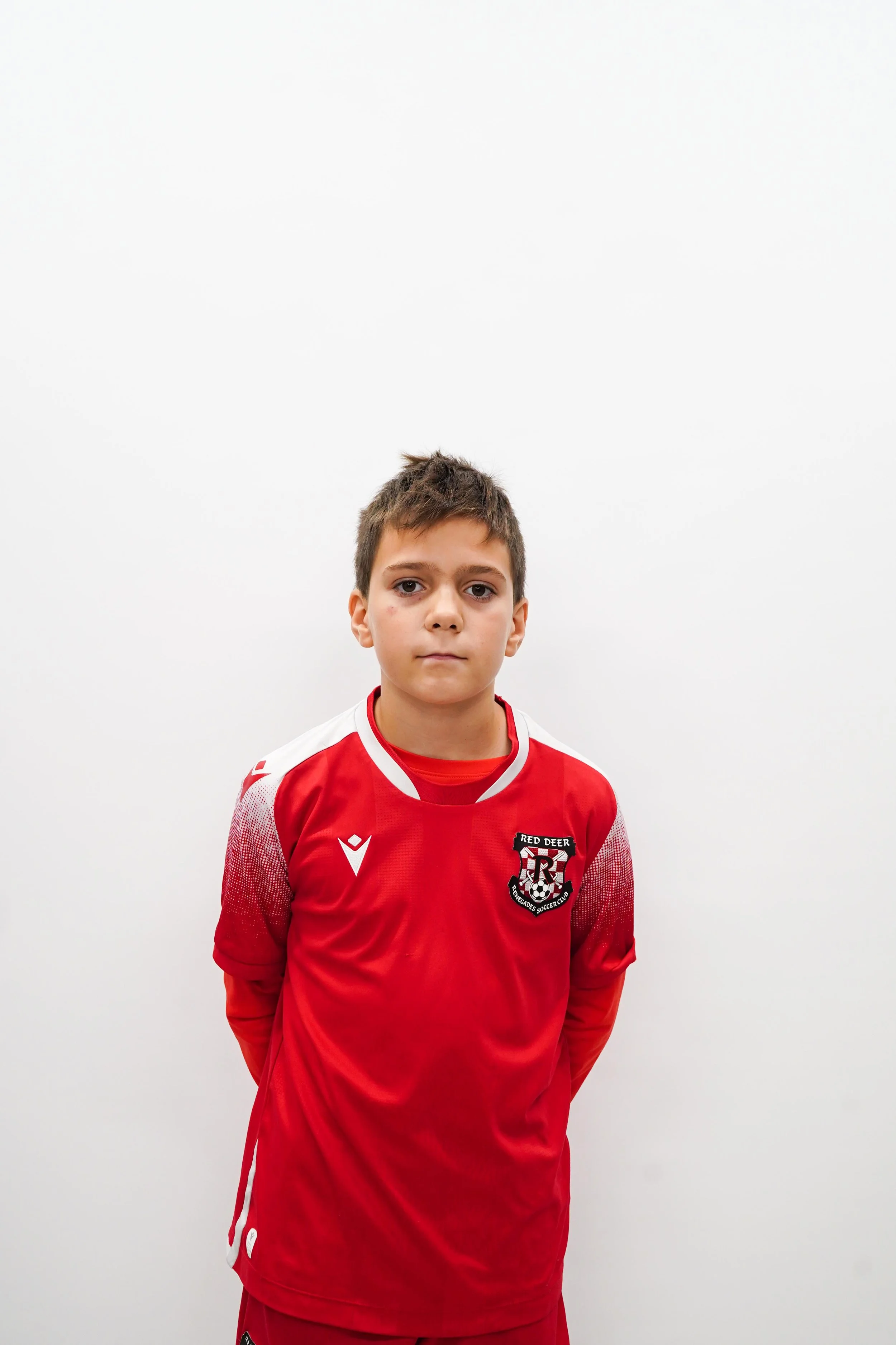 Young boy wearing a red soccer uniform with a crest that says 'Red Deer' standing against a plain white background.