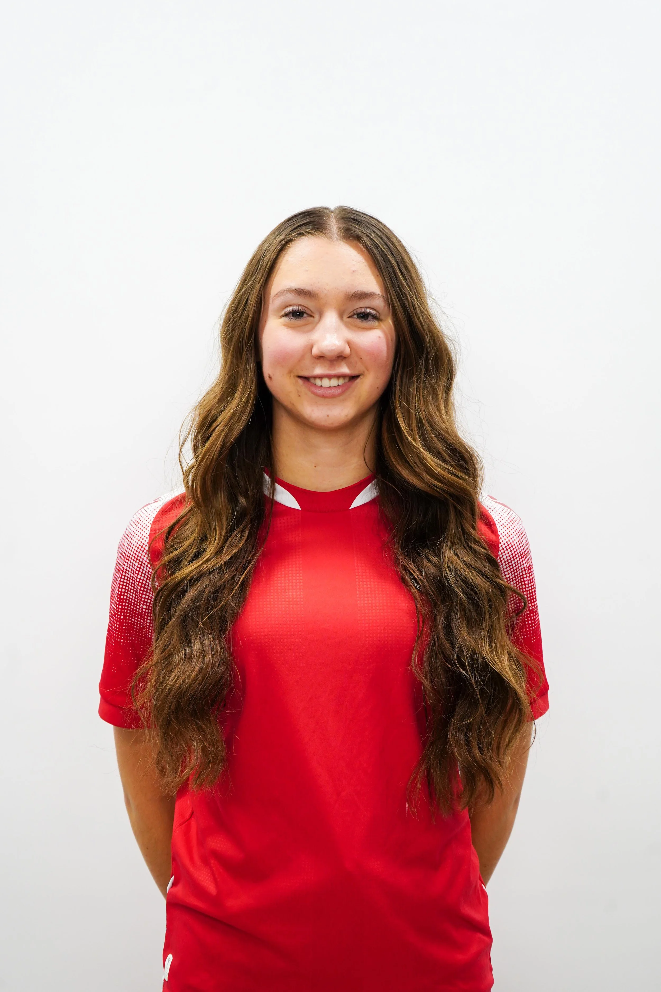 Young woman with long wavy brown hair smiling, wearing a red sports jersey, standing against a plain white background.