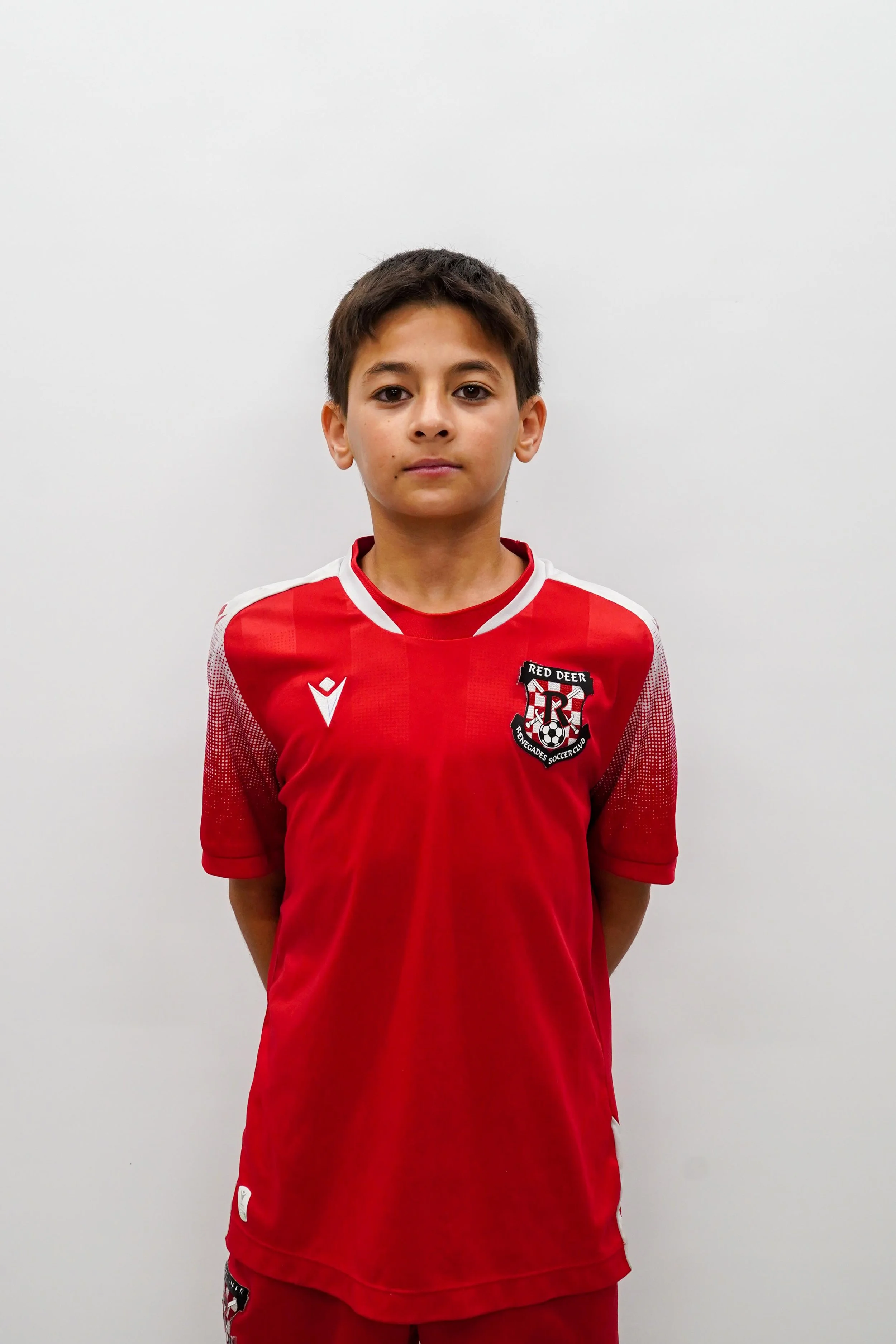 A young boy wearing a red soccer uniform with a crest on the chest, standing against a plain white wall.