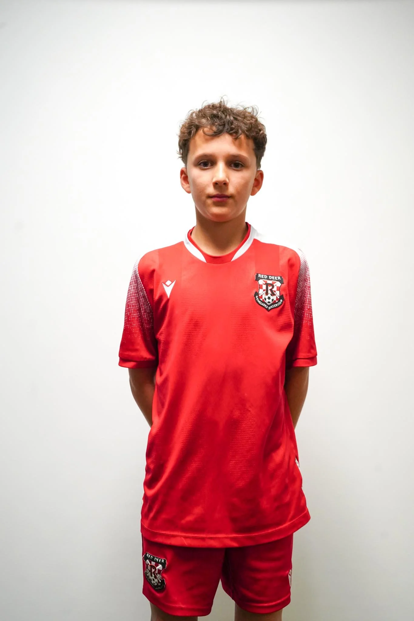 Young boy wearing a red soccer uniform with the Red Deer Soccer Club logo, standing against a plain white wall.